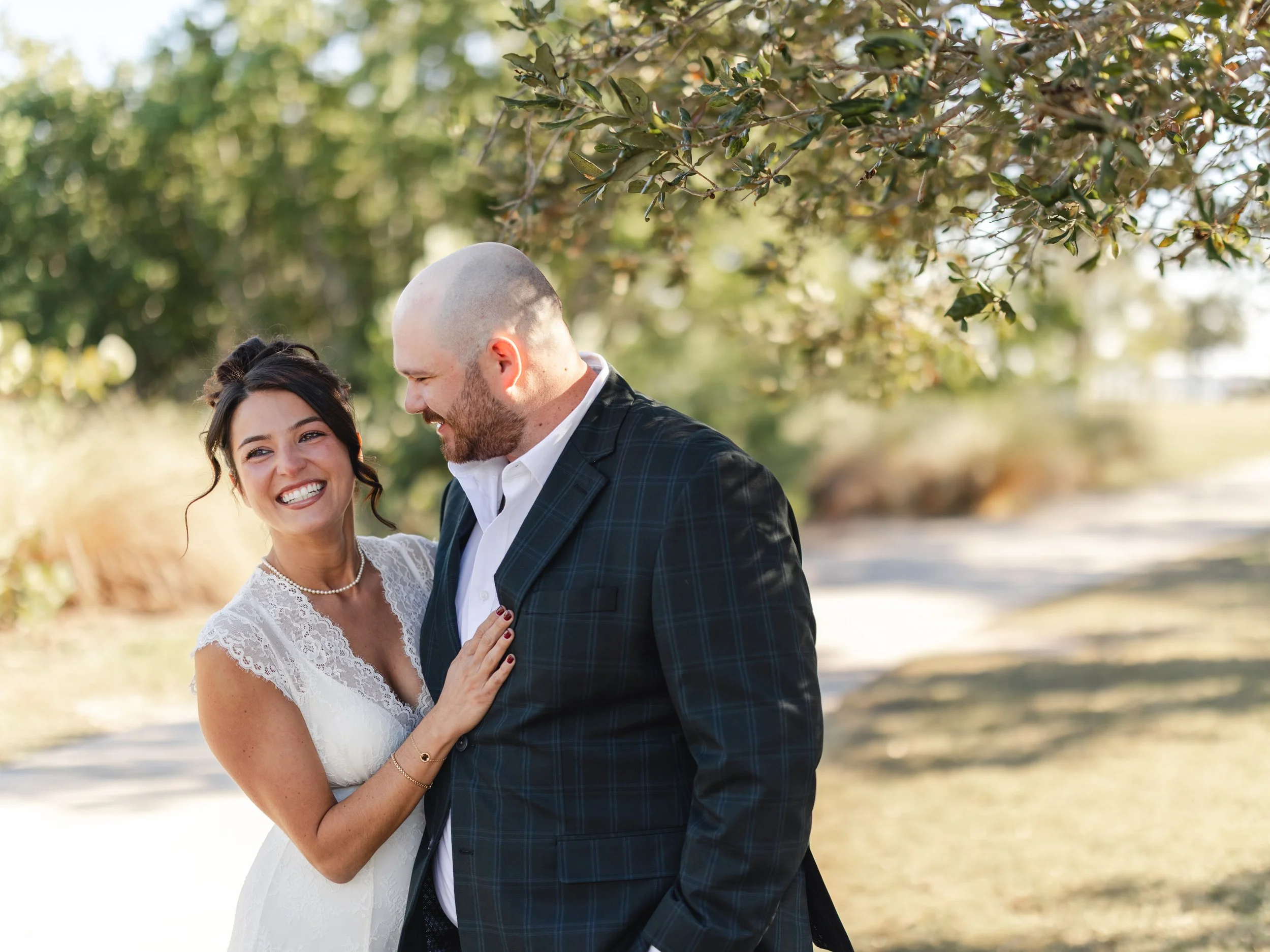 A smiling woman in a white lace wedding dress standing close to a man in a dark plaid suit outdoors with trees in the background.