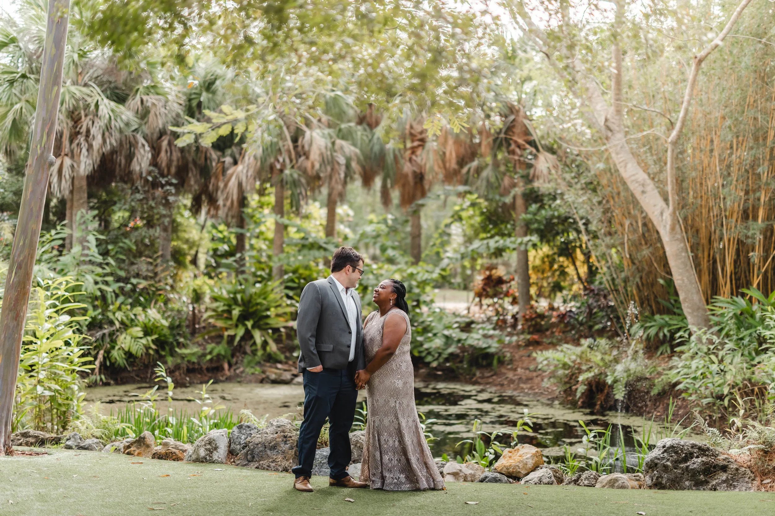 A couple dressed in formal attire standing close together by a pond surrounded by lush greenery and trees in a park.