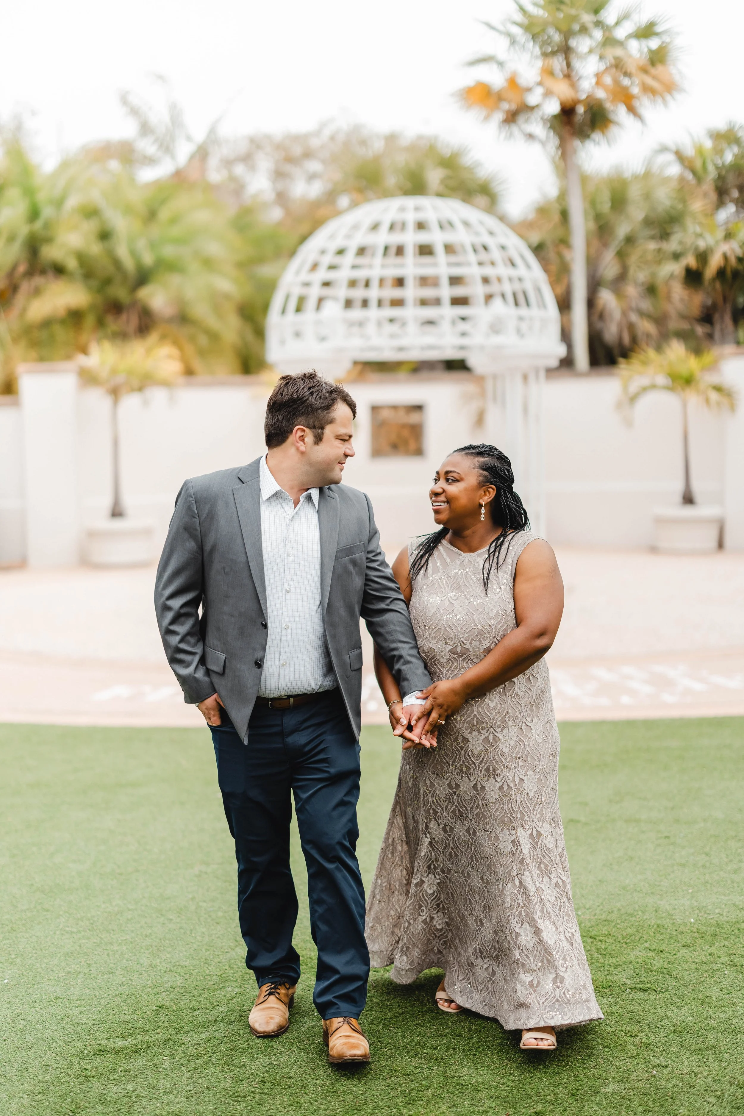 A couple holding hands, smiling at each other, outdoors with greenery and a white architectural structure in the background.