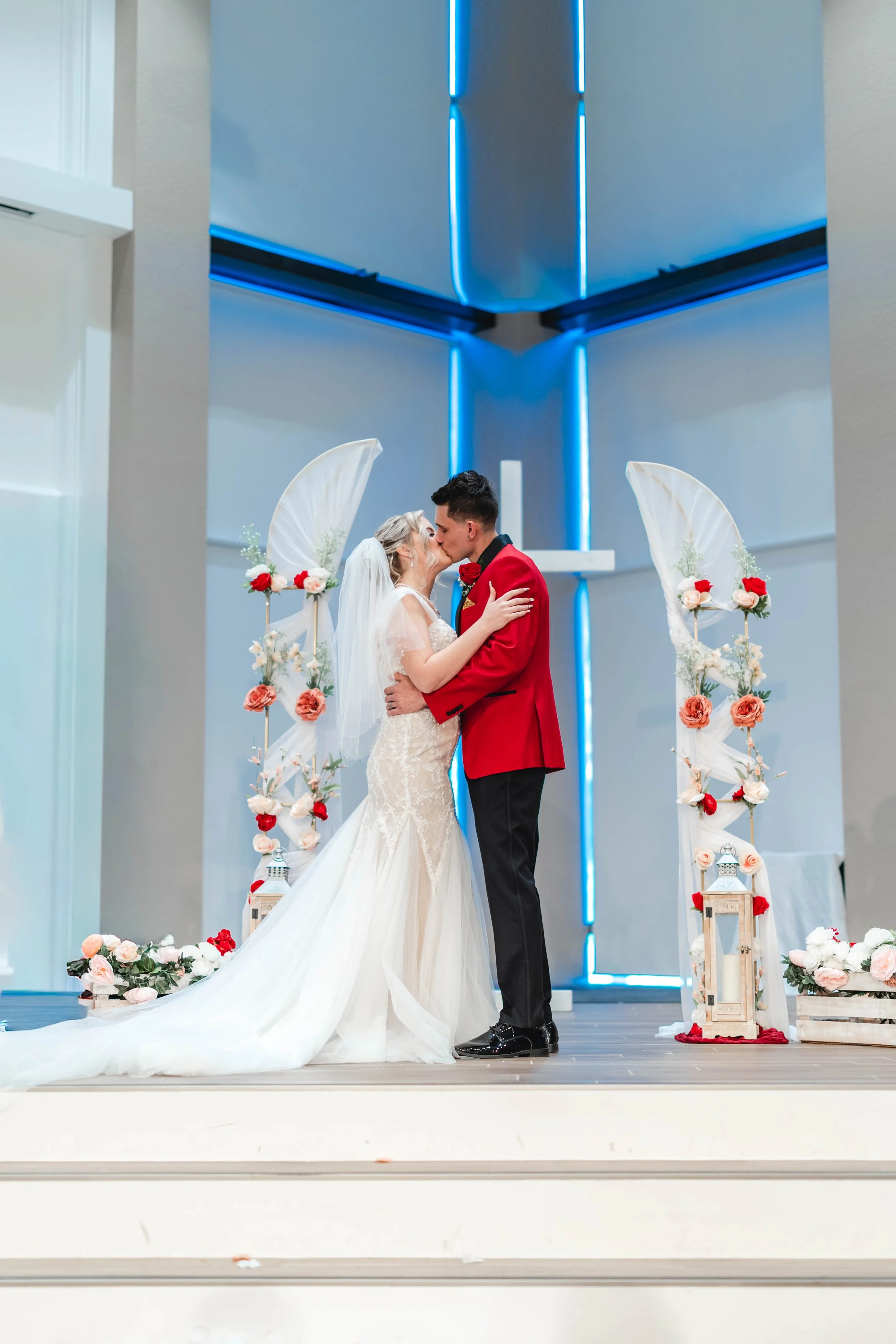 Bride and groom sharing a kiss during their wedding ceremony in a modern church with blue neon lighting and floral decorations.