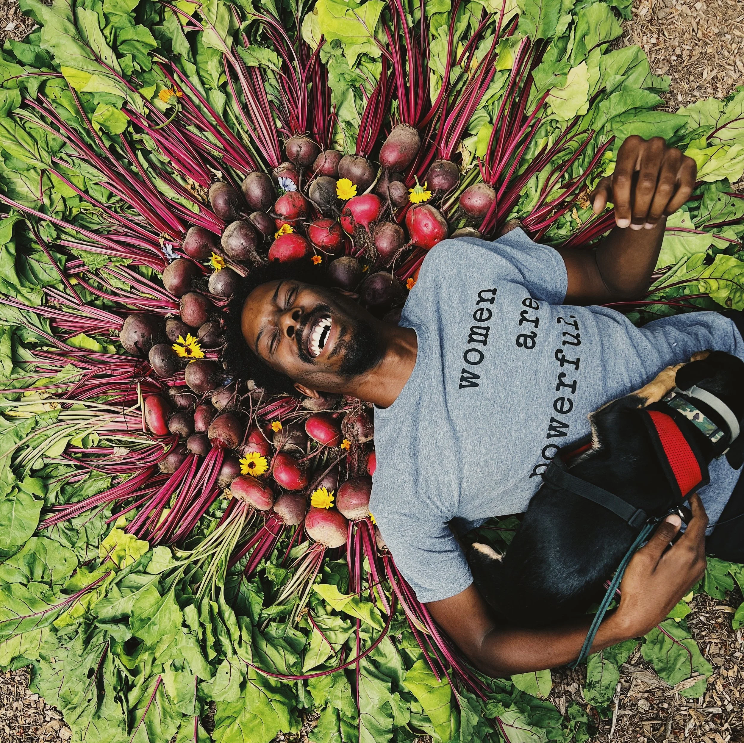 Person with a big smile laying down on top of a collection of beautiful vegetables and flowers