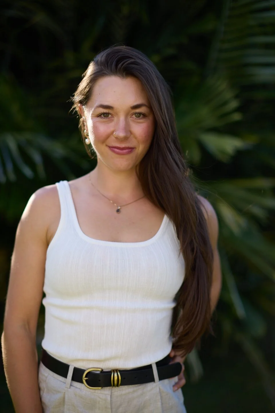 A young woman with long dark hair, smiling, wearing a white sleeveless top and black belt, standing outdoors with greenery in the background.