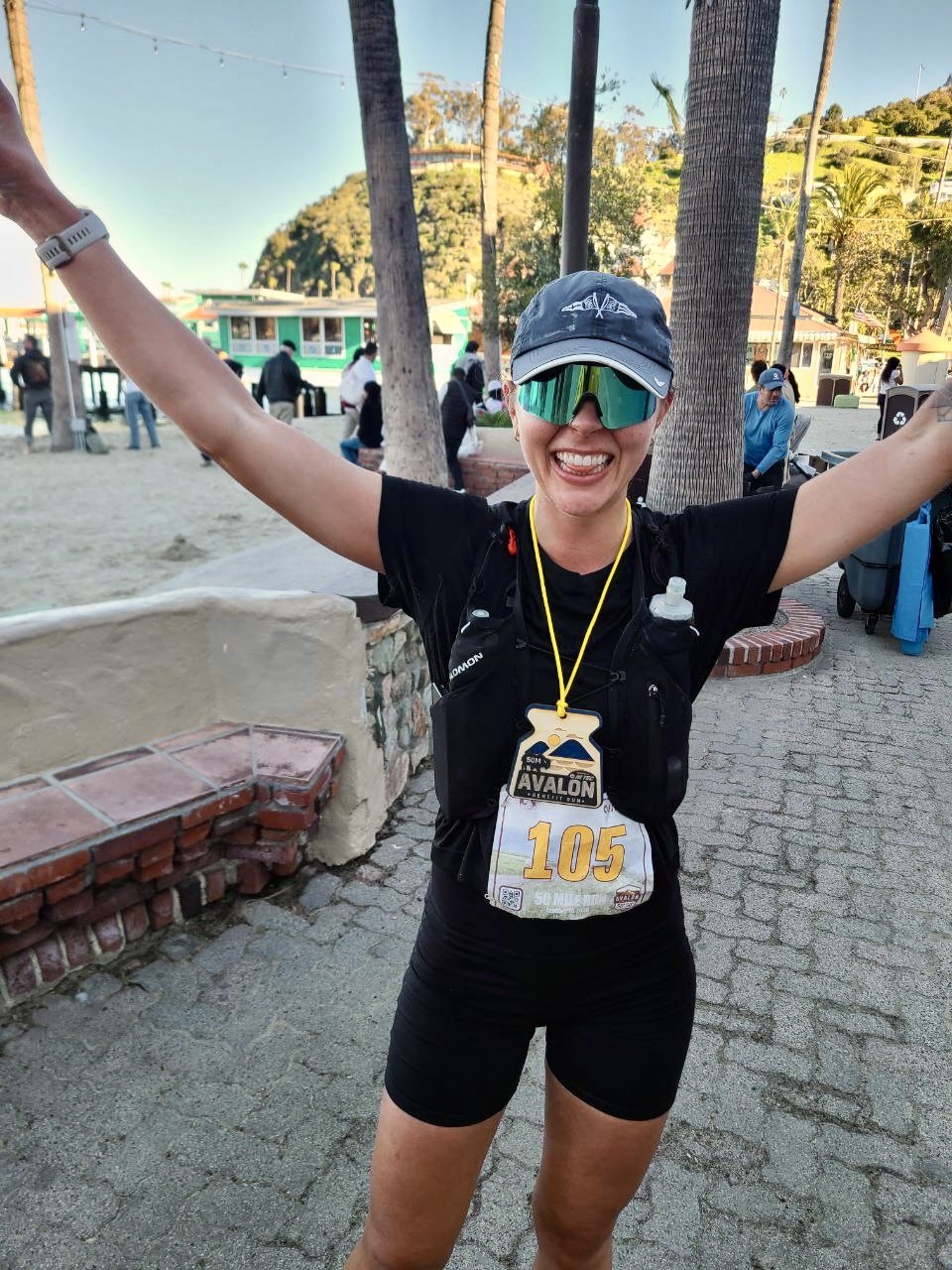 A woman with a big smile, wearing sunglasses, a cap, black running outfit, and a race bib with the number 105, celebrating running a 50 mile ultramarathon on Catalina island.