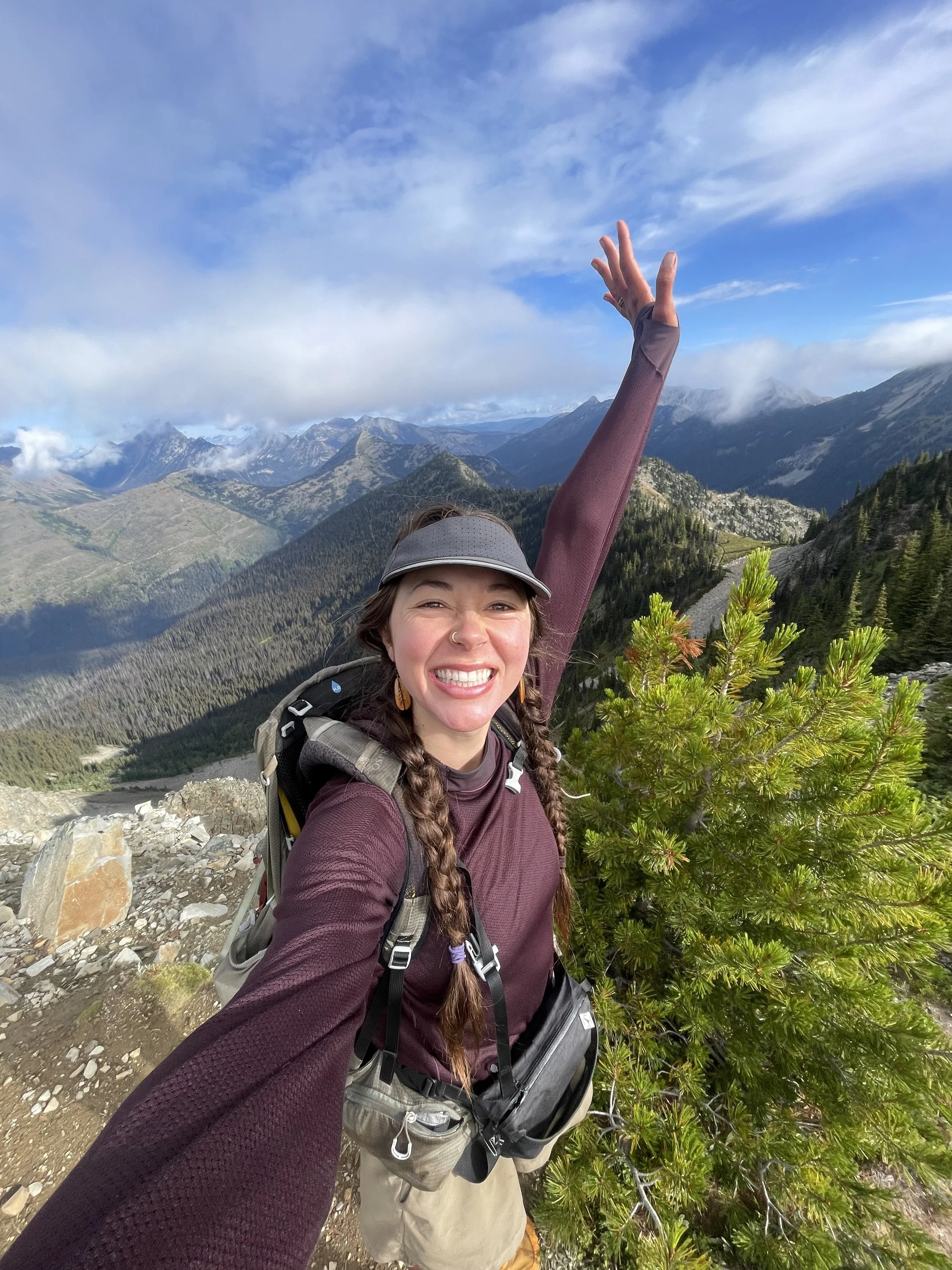 A woman smiling and celebrating on a mountain trail with a scenic view of mountains, forests, and a partly cloudy sky in the background.