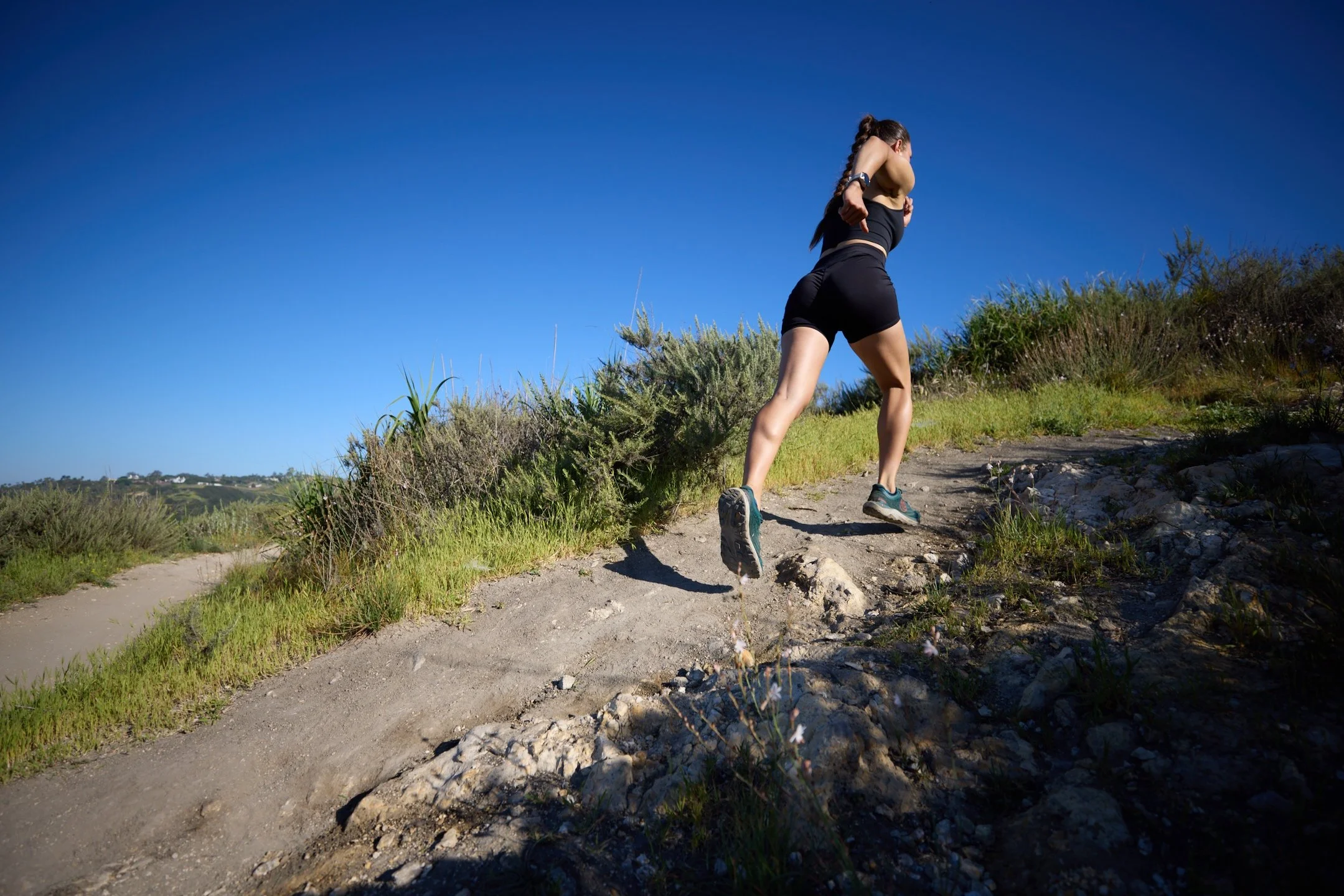 Woman running uphill on a dirt trail surrounded by grass and low shrubs under a clear blue sky.