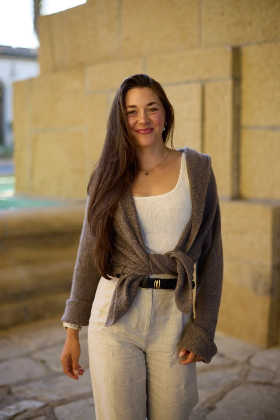 A young woman with long brown hair wearing a white top, beige pants, and a tied brown cardigan, standing outdoors near a beige stone wall, smiling at the camera.