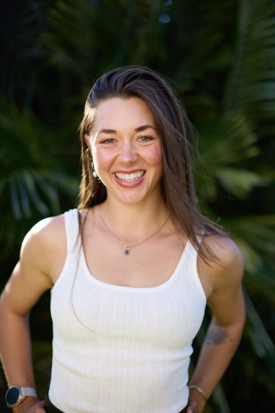 A young woman smiling outdoors, wearing a white tank top, jewelry, and a smartwatch, with green tropical plants in the background.