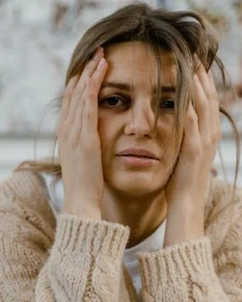 A woman with brown hair looking distressed, holding her head with both hands, wearing a beige knit sweater.