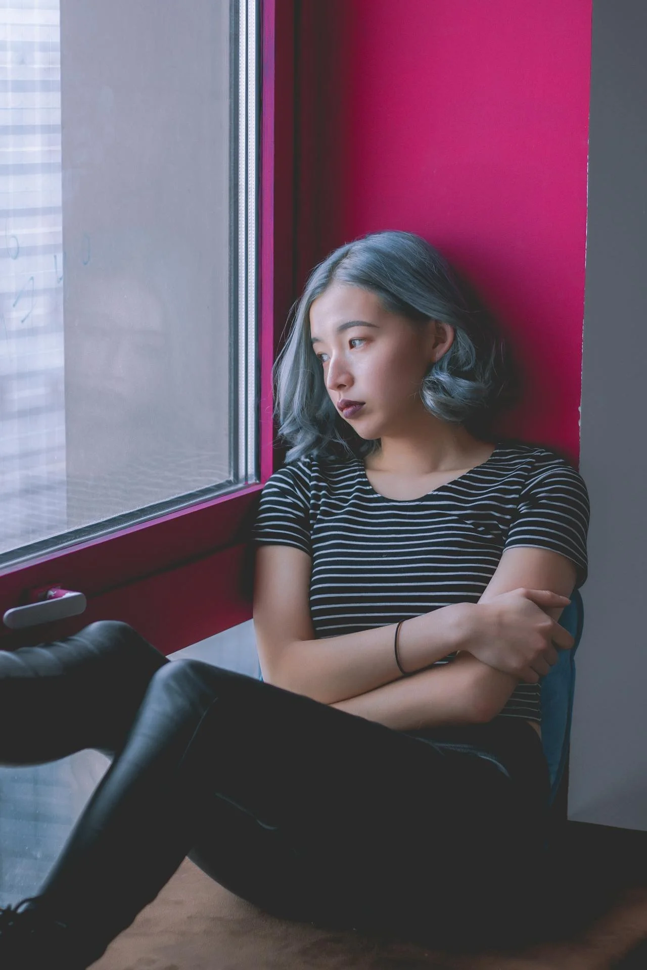 A woman sits on the floor and looks glumly out a window.