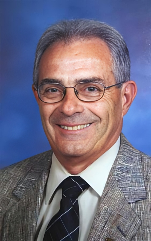 A middle-aged man with short gray hair wearing glasses, a plaid suit jacket, white shirt, and a black tie, smiling against a blue background.