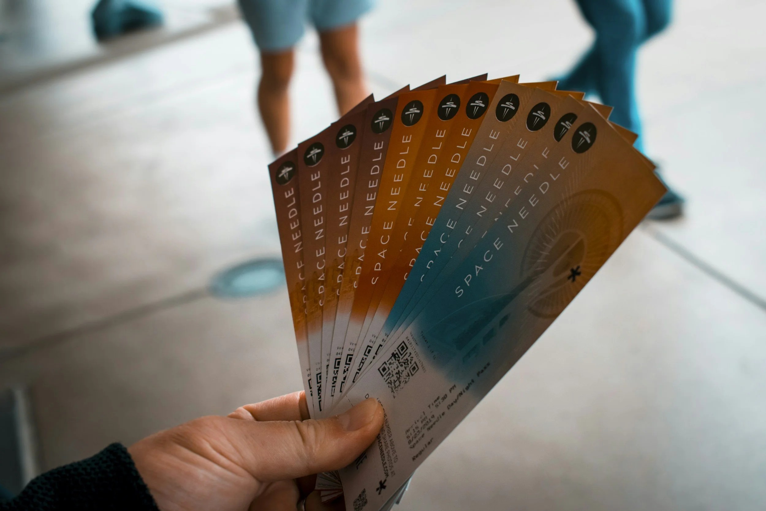 Person holding a fan of orange and blue airline boarding passes with 'SPACE NEEDLE' printed on them, others with QR codes visible in background.