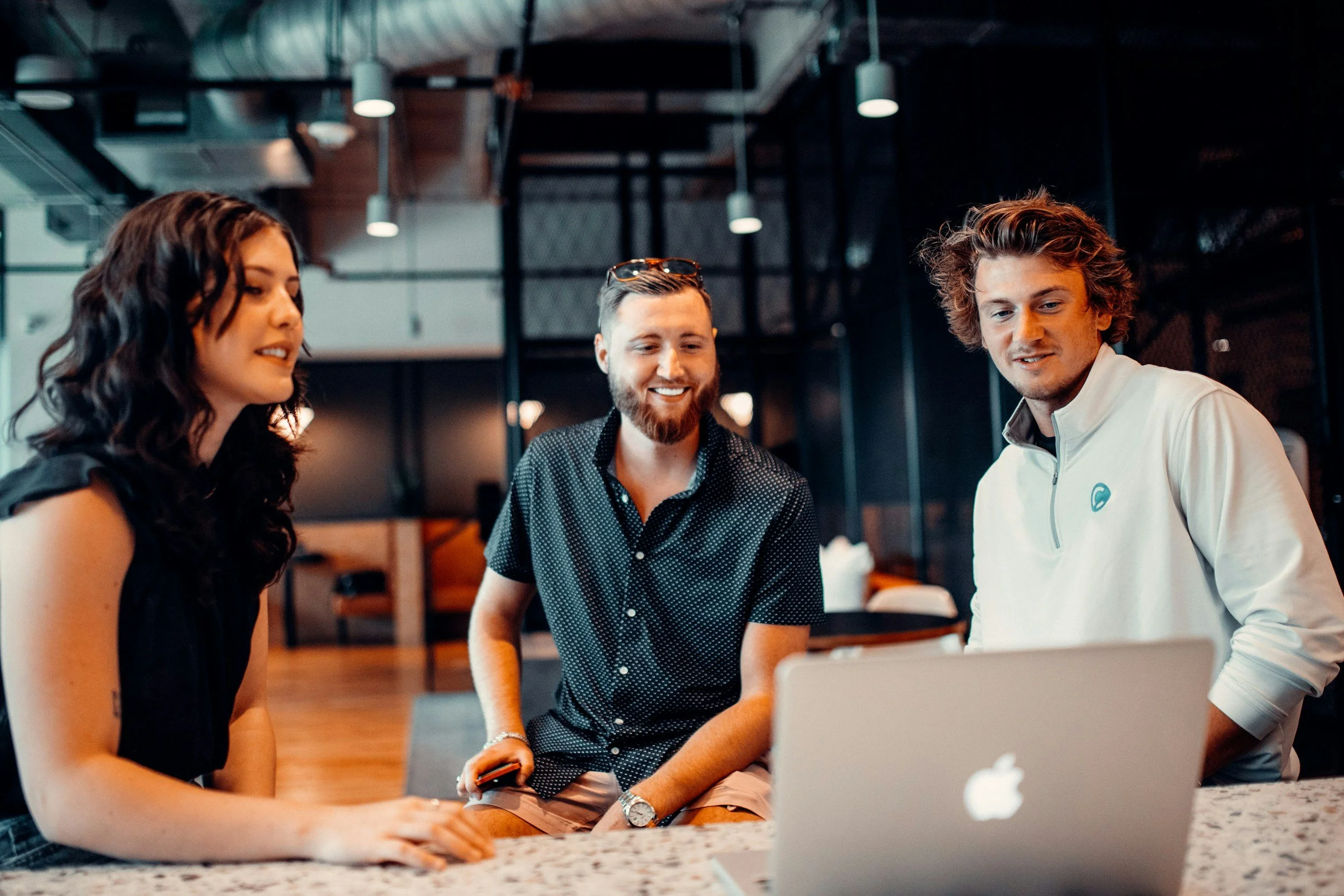 Three young adults gathered around a laptop in a modern office or coworking space, engaging in a discussion and smiling.