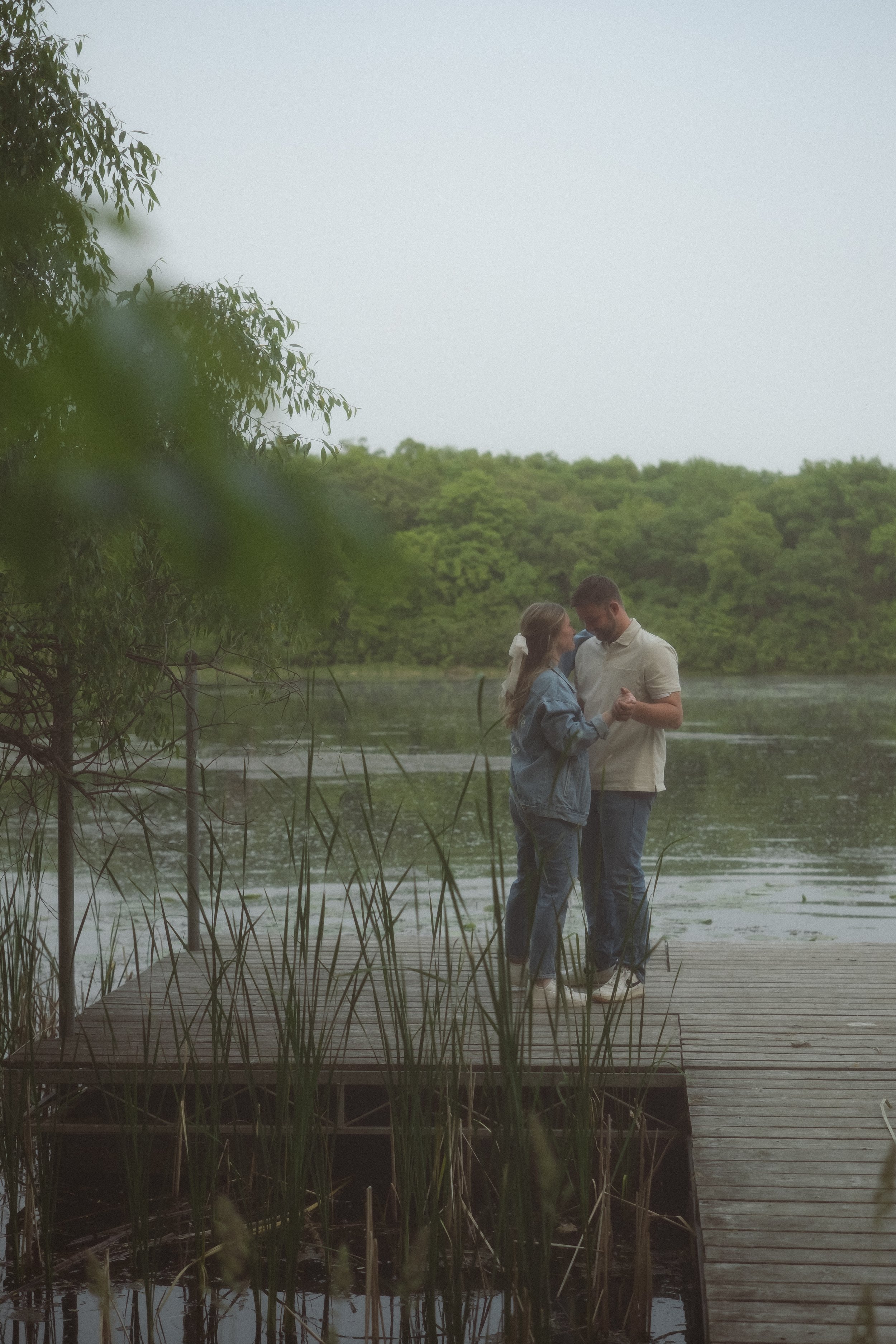 A couple dancing on a wooden dock near a lake with green trees in the background.
