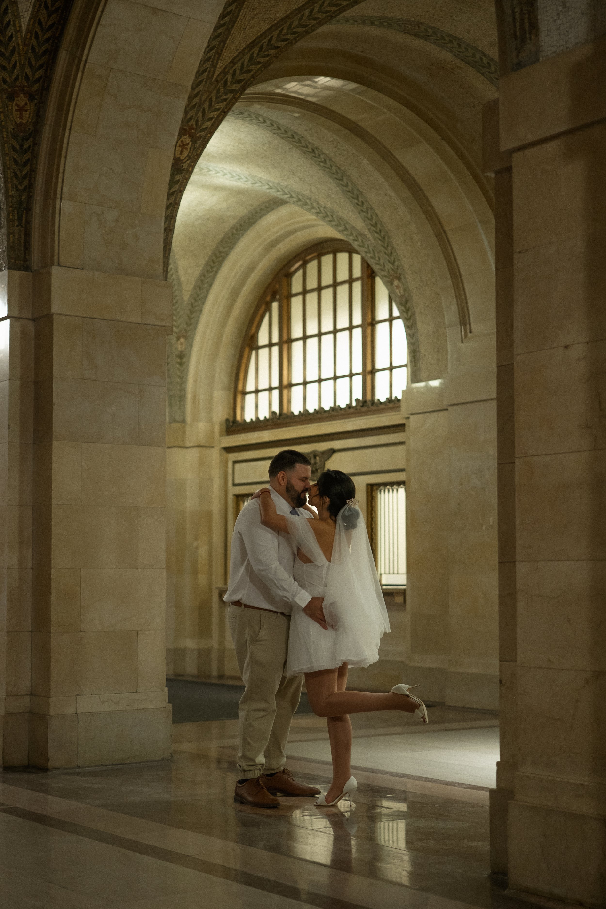 A couple in wedding attire embracing inside a grand, ornate building with tall arched windows and detailed ceiling architecture.