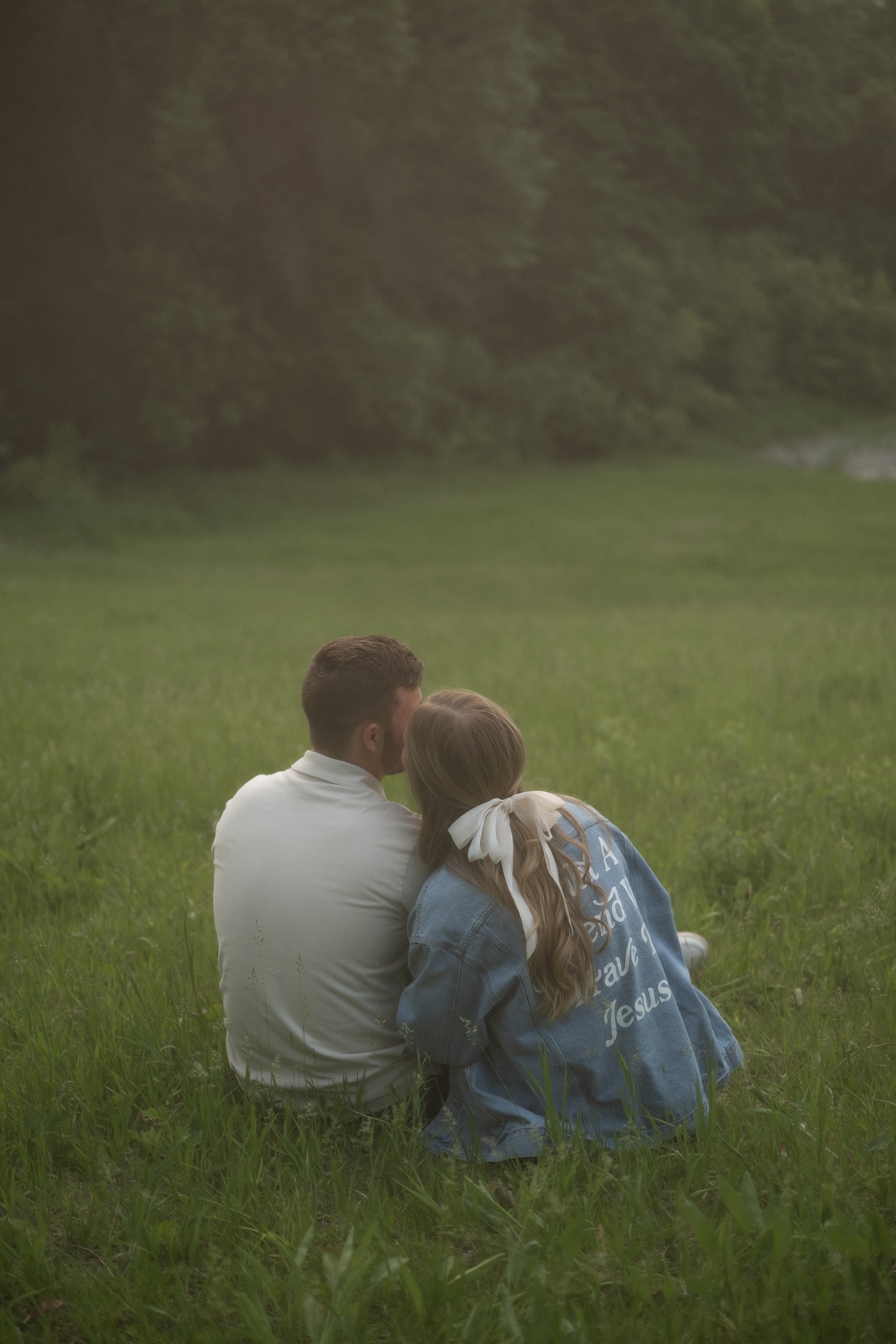 A young couple sitting on the grass in a field, with the woman leaning her head on the man's shoulder, both facing away from the camera, surrounded by greenery and trees.