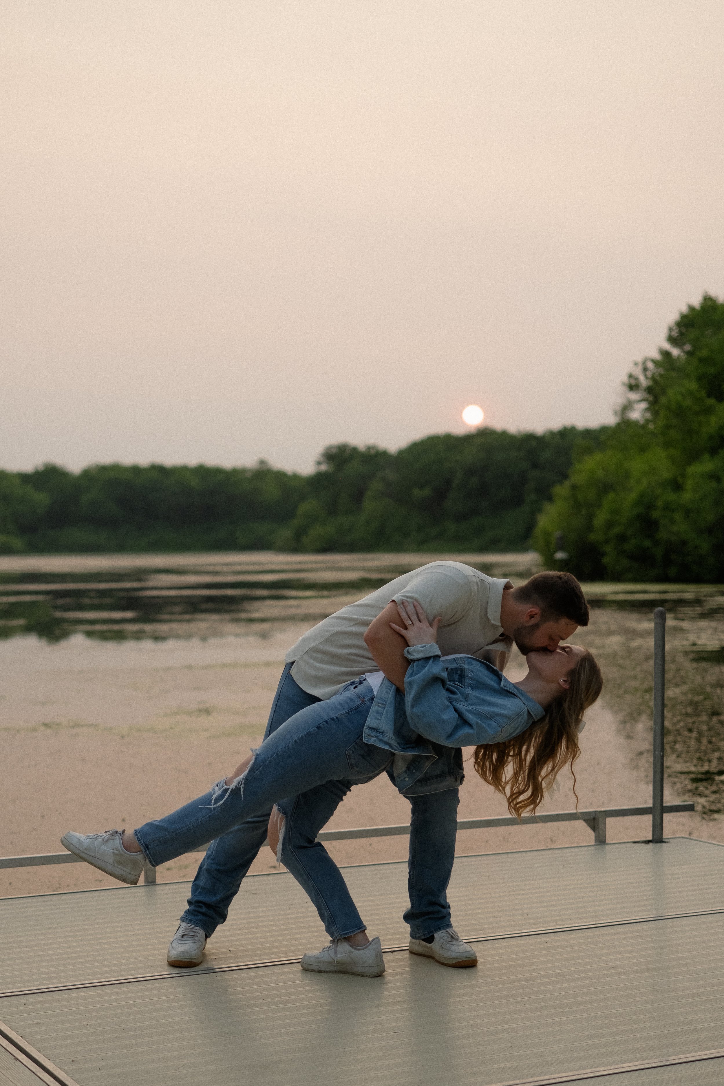A couple is sharing a kiss on a dock at sunset by a lake, with the man dipping the woman backwards. The scenery includes water, green trees, and a setting sun in the background.
