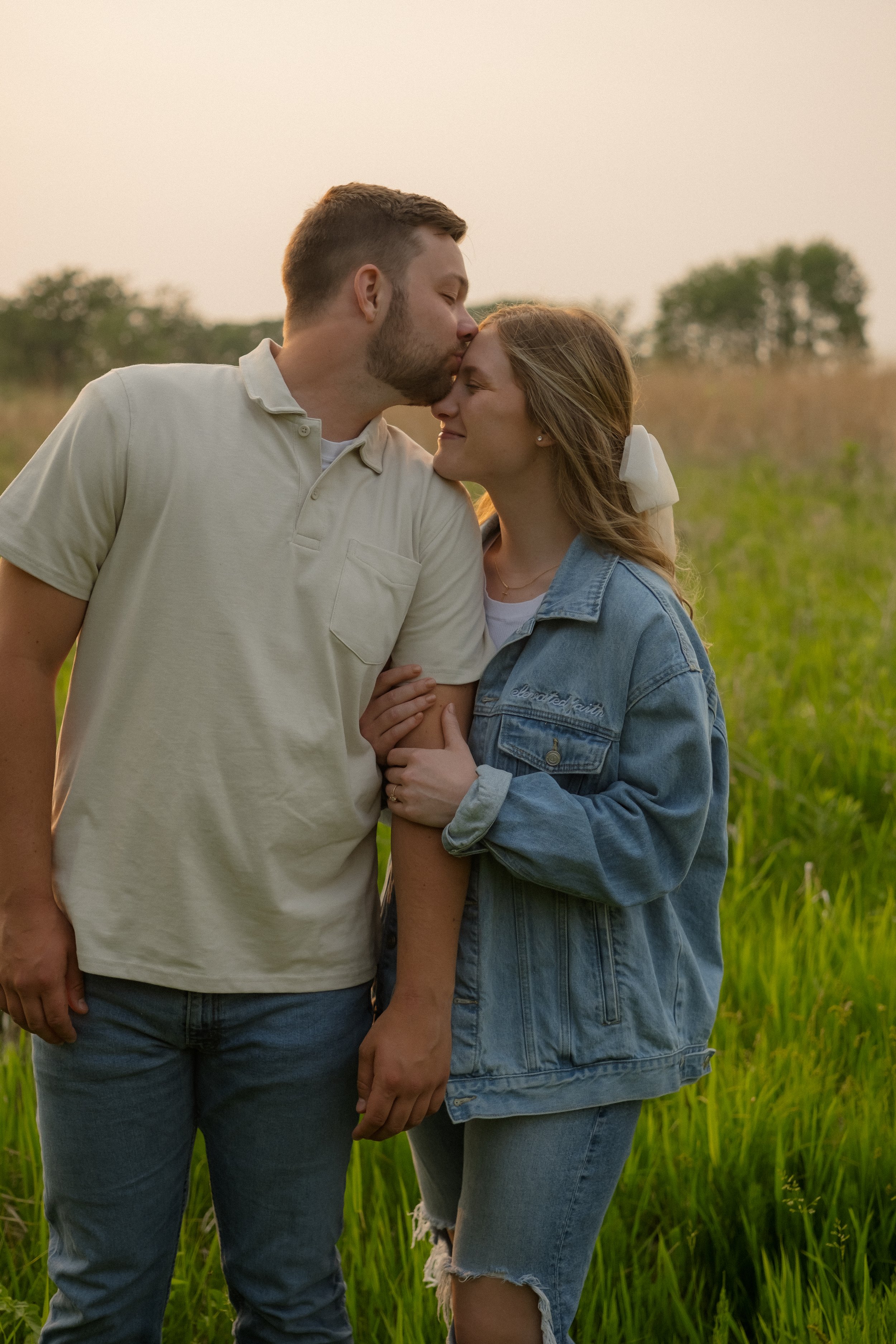 A couple standing in a grassy field during sunset, with the man kissing the woman's forehead.