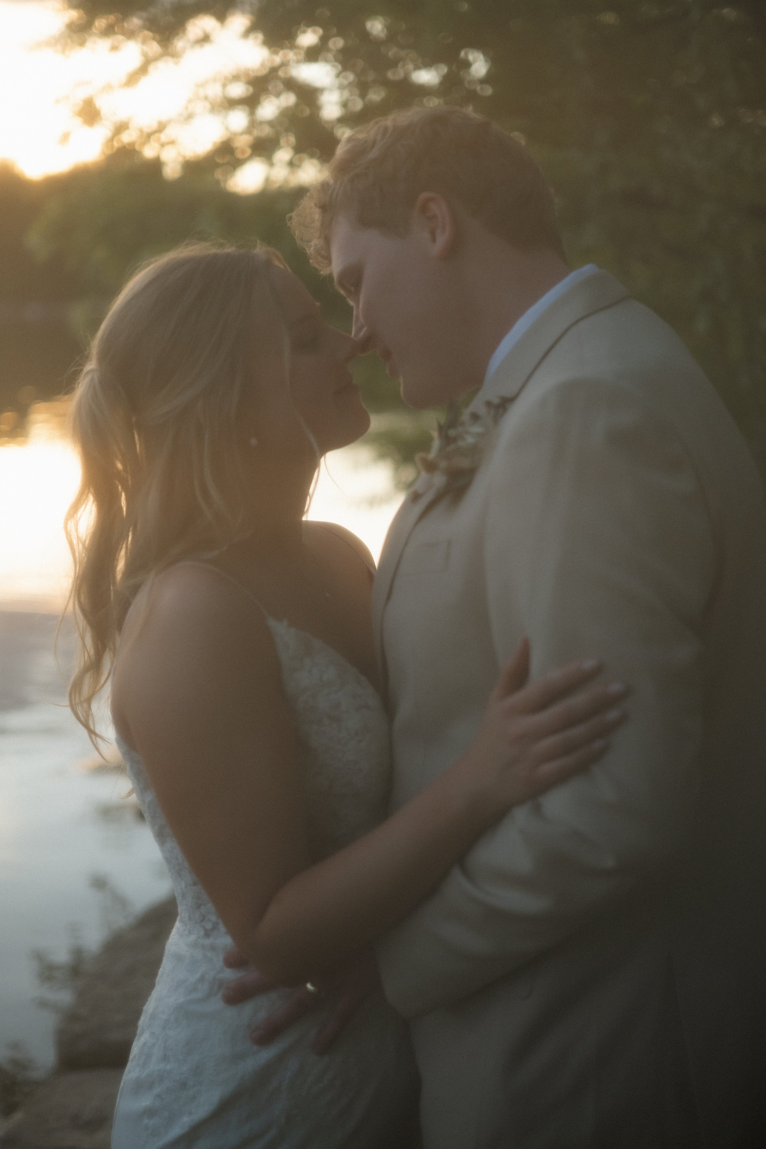 A romantic couple, dressed in wedding attire, sharing an intimate moment in front of a sunset by the water.