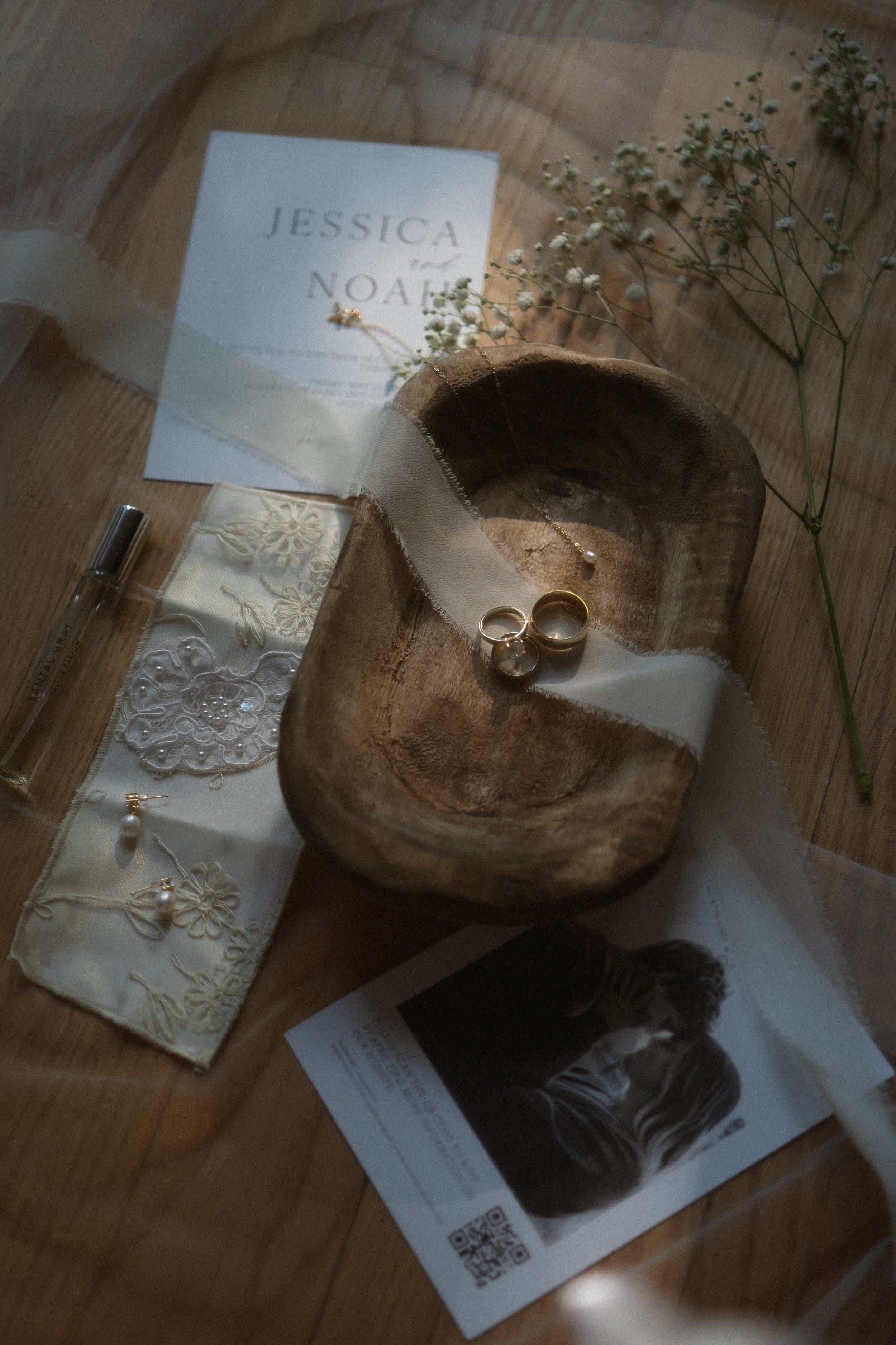 A wooden bowl holding two wedding rings and a pearl necklace, surrounded by lace fabric, a wedding invitation for Jessica and Noah, a black and white photo, and sprigs of baby's breath flowers on a wooden surface.