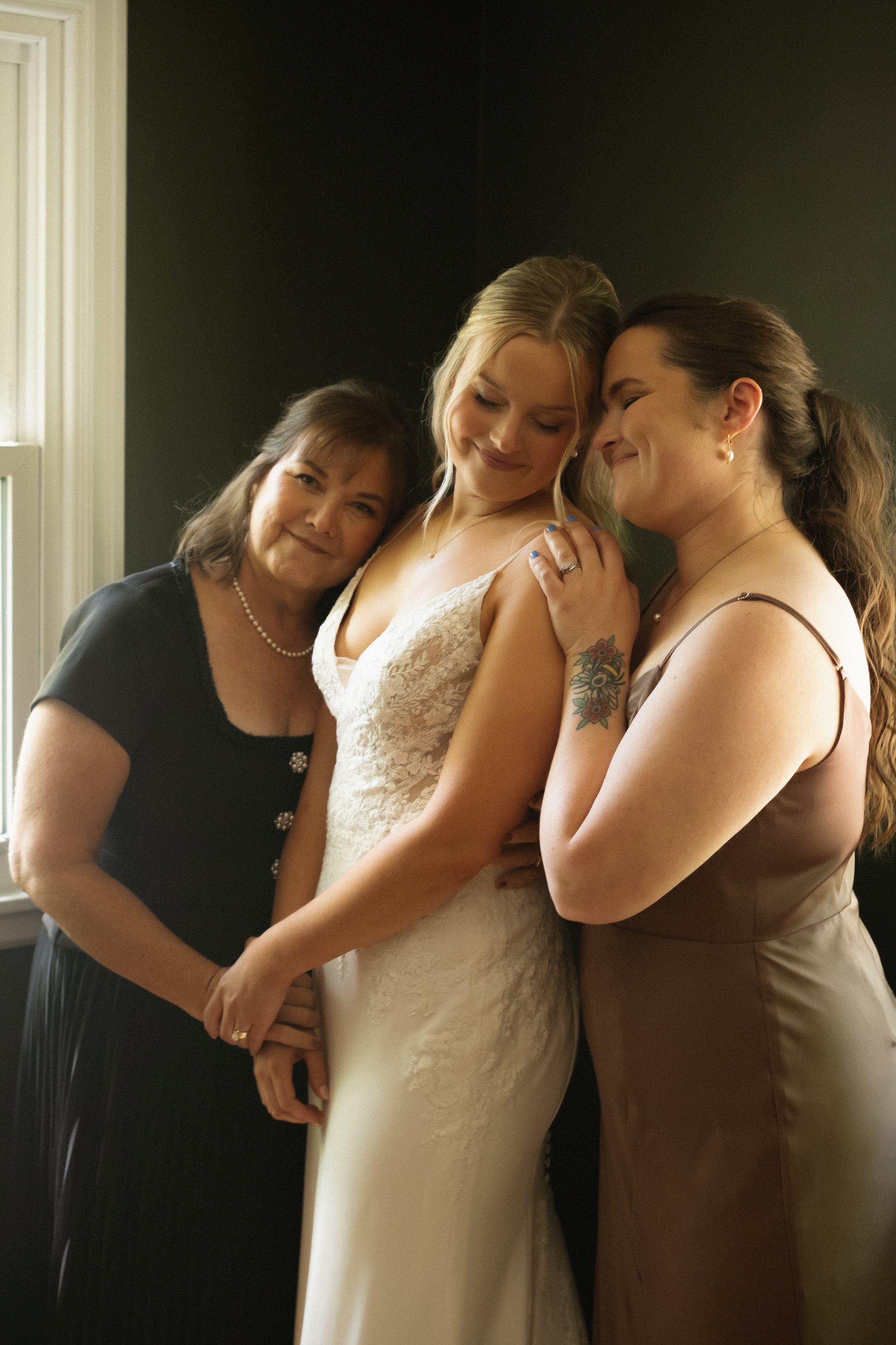 Three women, including a bride in a white wedding dress, embracing and smiling together indoors near a window.