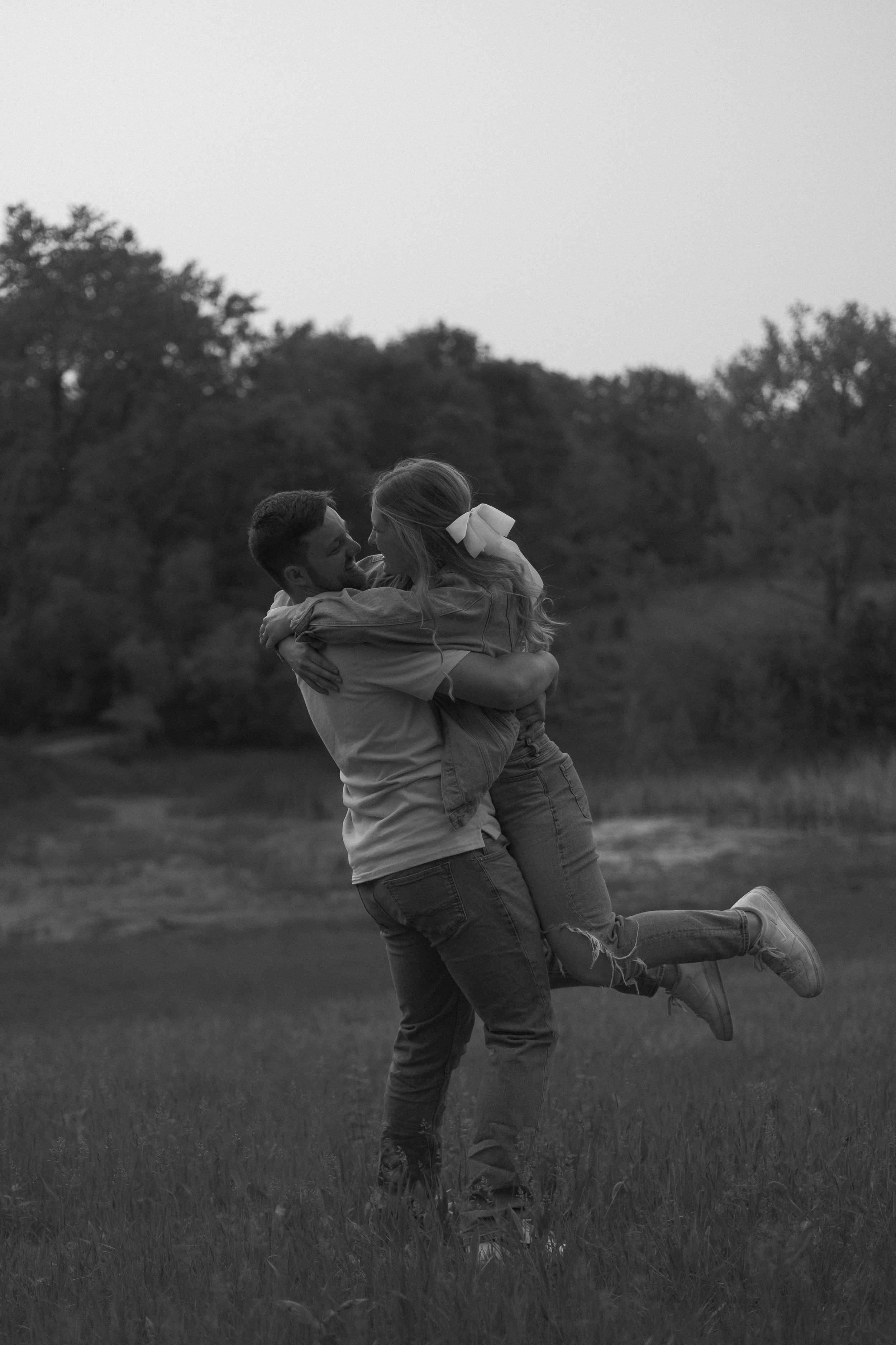 A couple is hugging and lifting each other in a grassy field with trees in the background, black and white photo.
