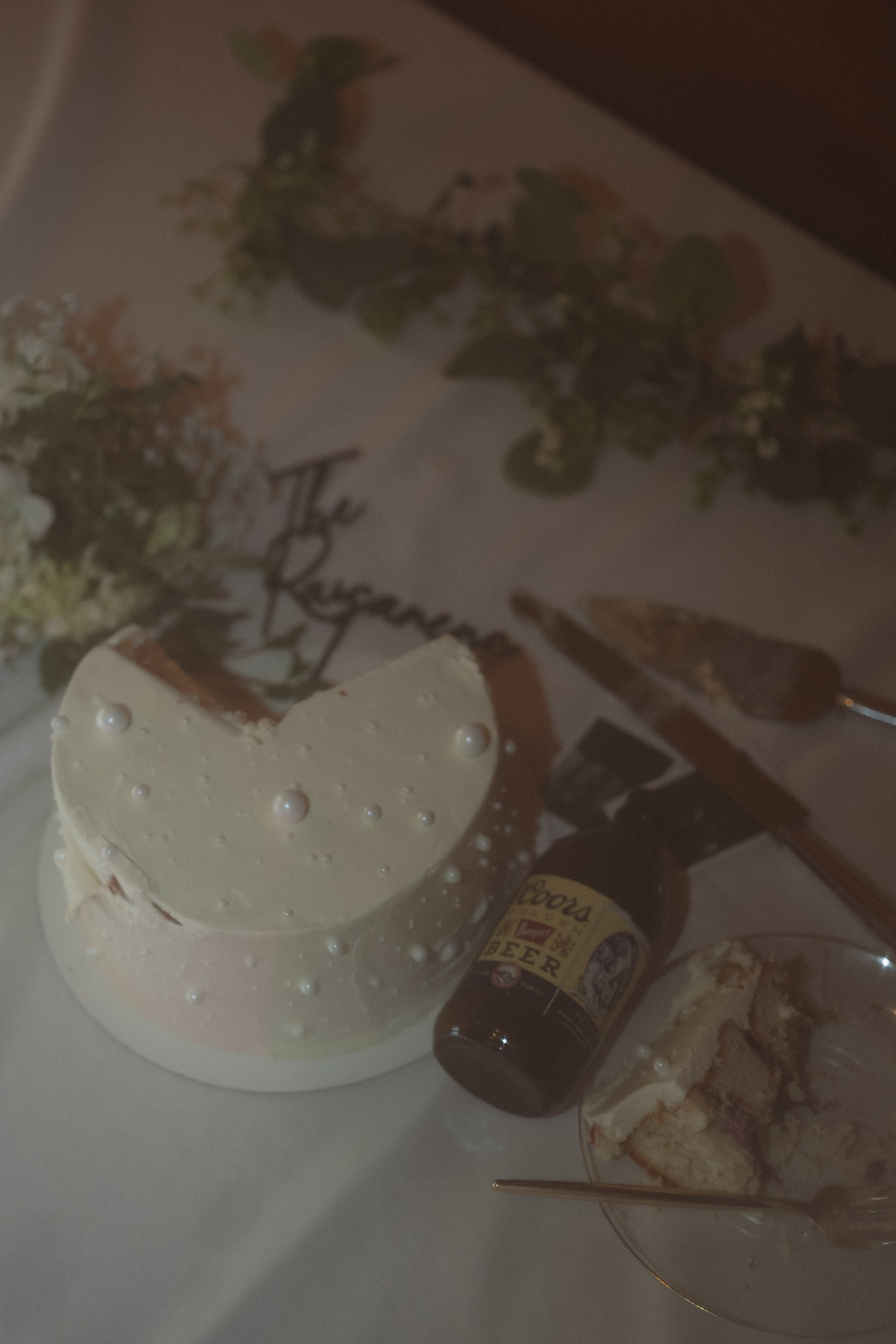 A birthday celebration setup featuring a white cake with pearl decorations, a glass of cake slices, a beer bottle, and a backdrop with floral decorations and text that reads 'The Ruggers.'