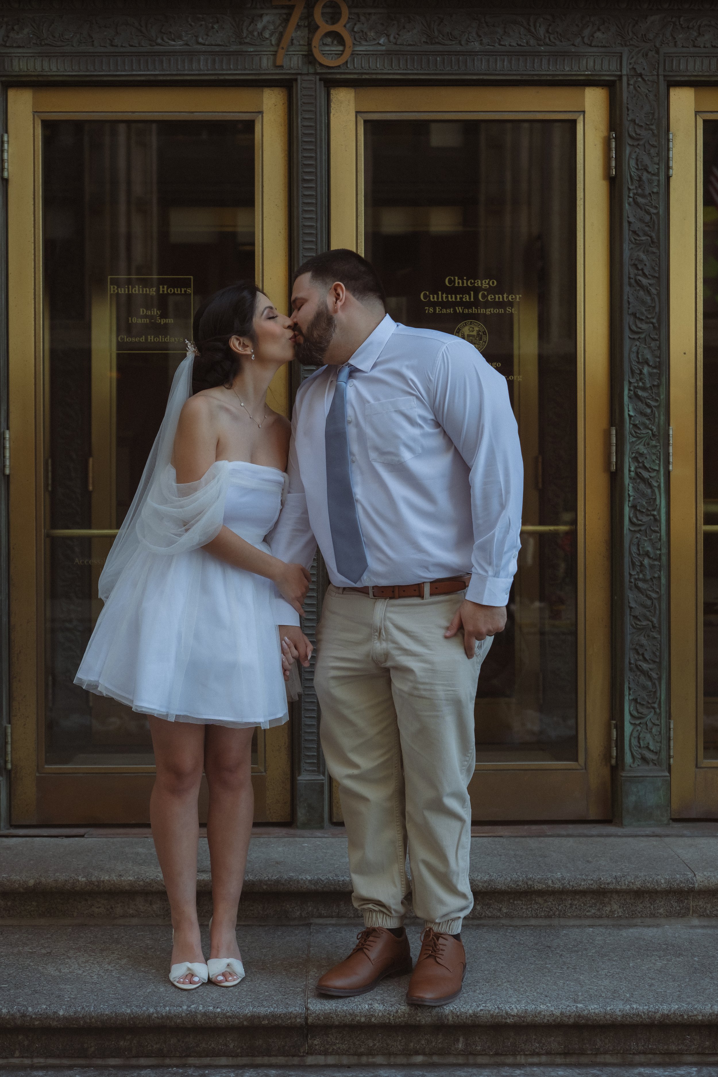 A bride and groom share a kiss outside a building with golden doors and ornate architectural details.