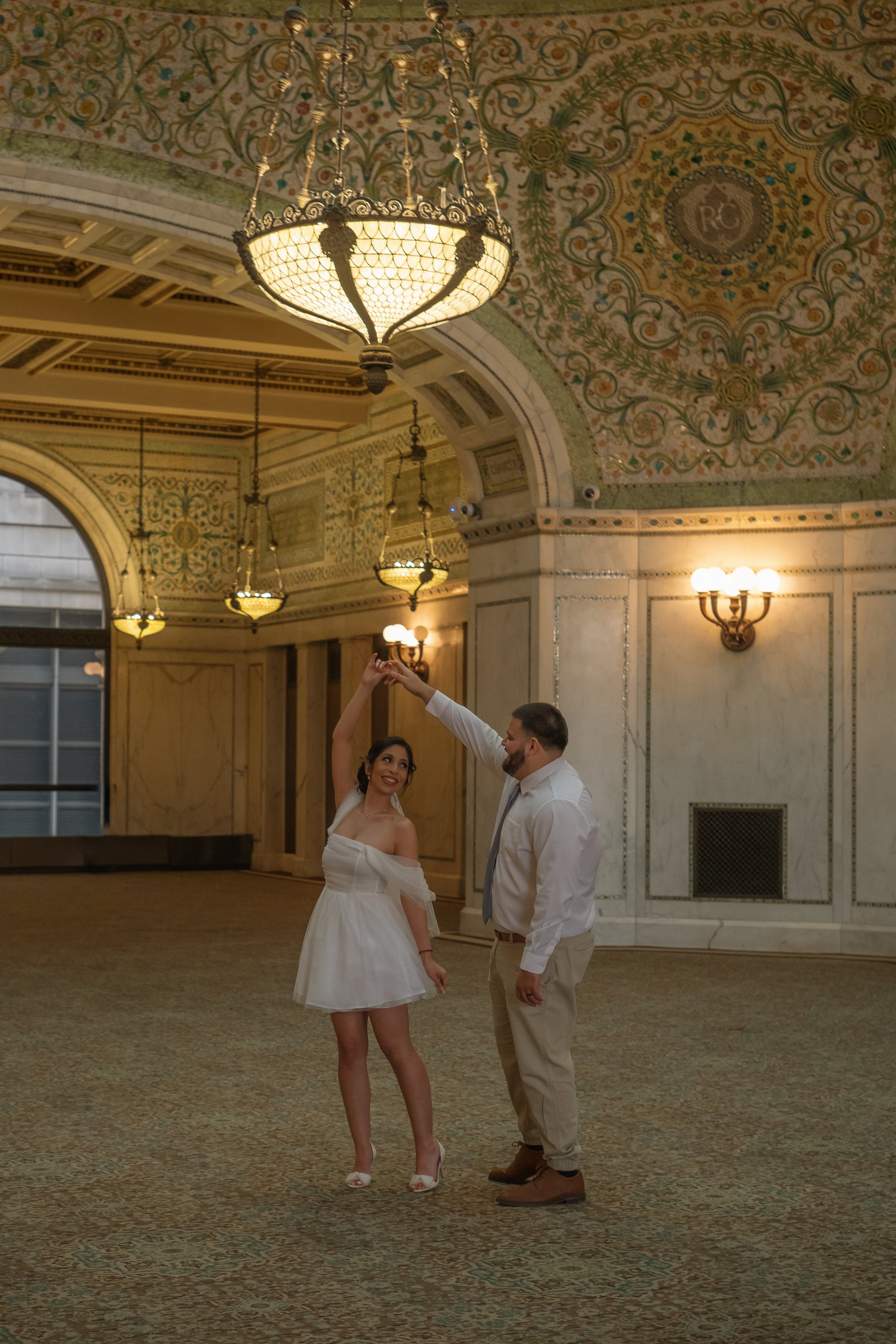 A woman in a white dress and a man in khaki pants and a white shirt dance together in a grand, ornate room with chandeliers and decorative ceiling art.
