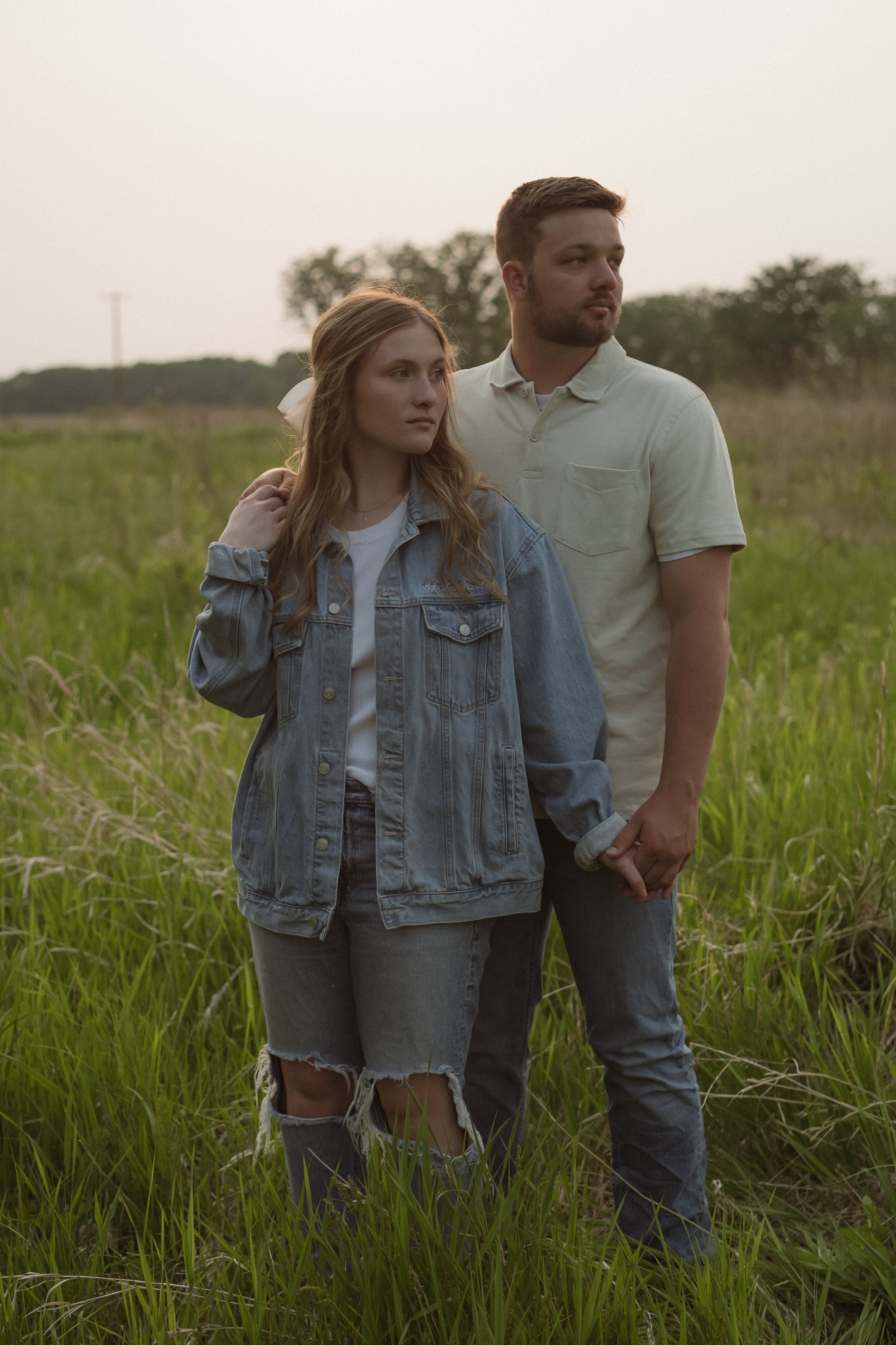 A young couple holding hands in a grassy field during sunset, with the woman wearing a denim jacket and ripped jeans, and the man dressed in a light-colored polo shirt and jeans.