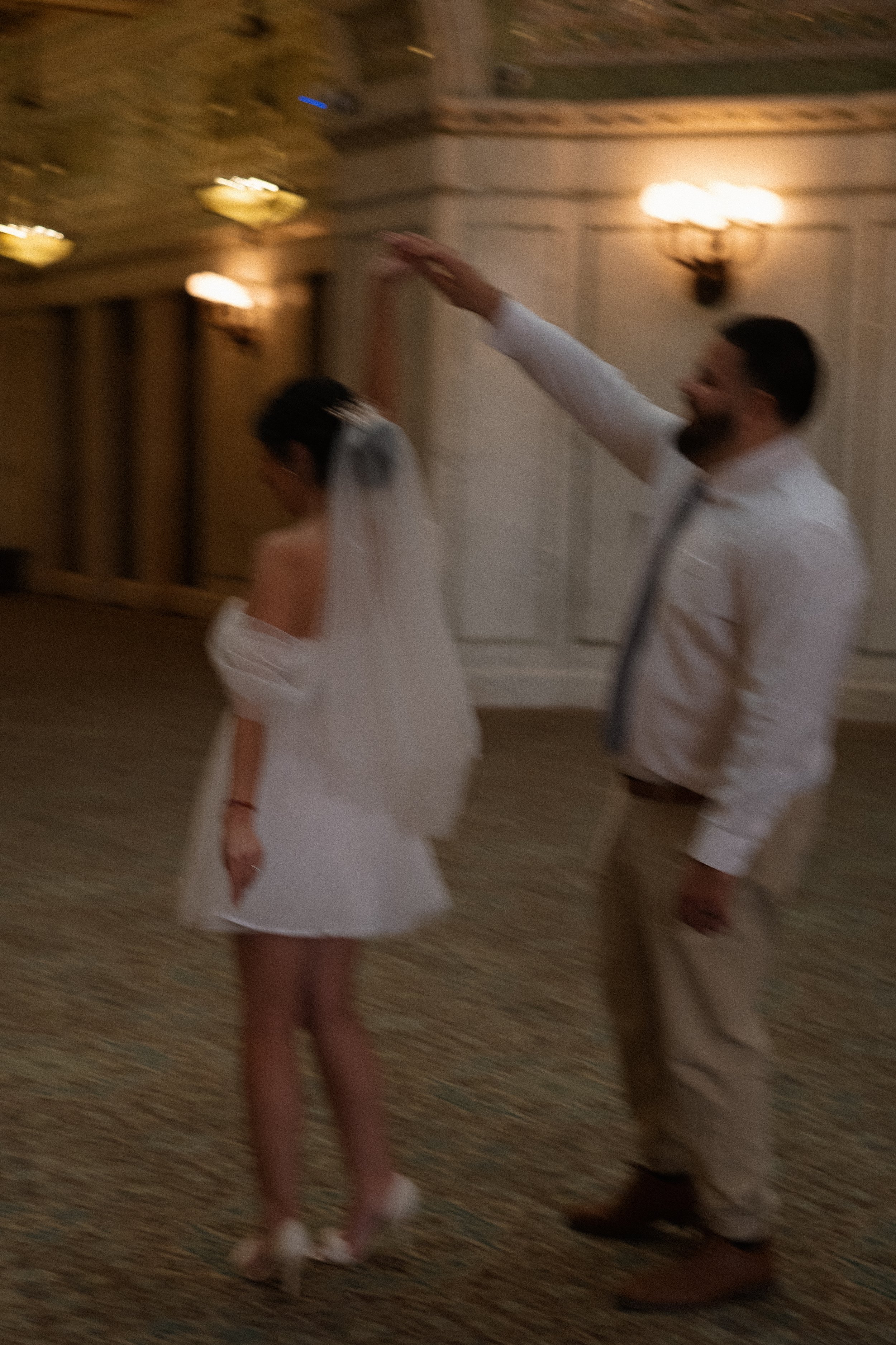 A man and woman dancing at a wedding reception, with the man spinning the woman. The woman is wearing a white dress and veil, and the man is dressed in a white shirt and beige pants.