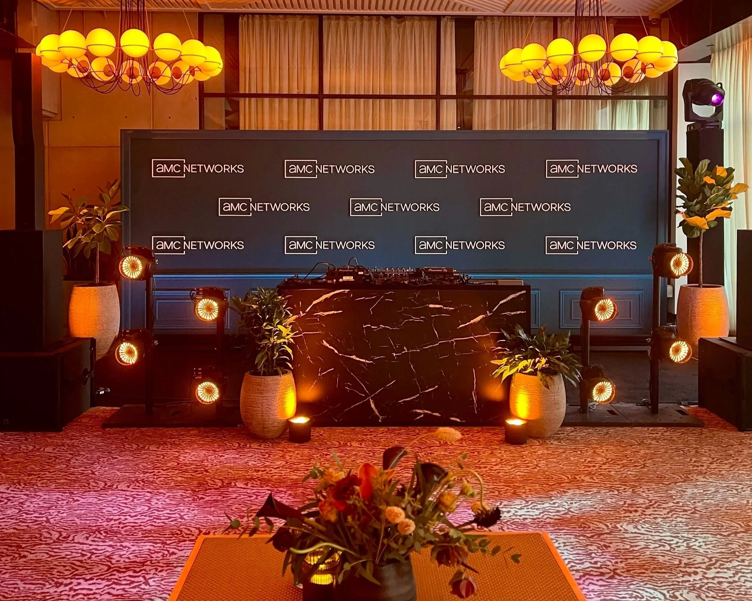 Step and repeat moment and DJ booth surrounded by lighting towers placed in front of wall.