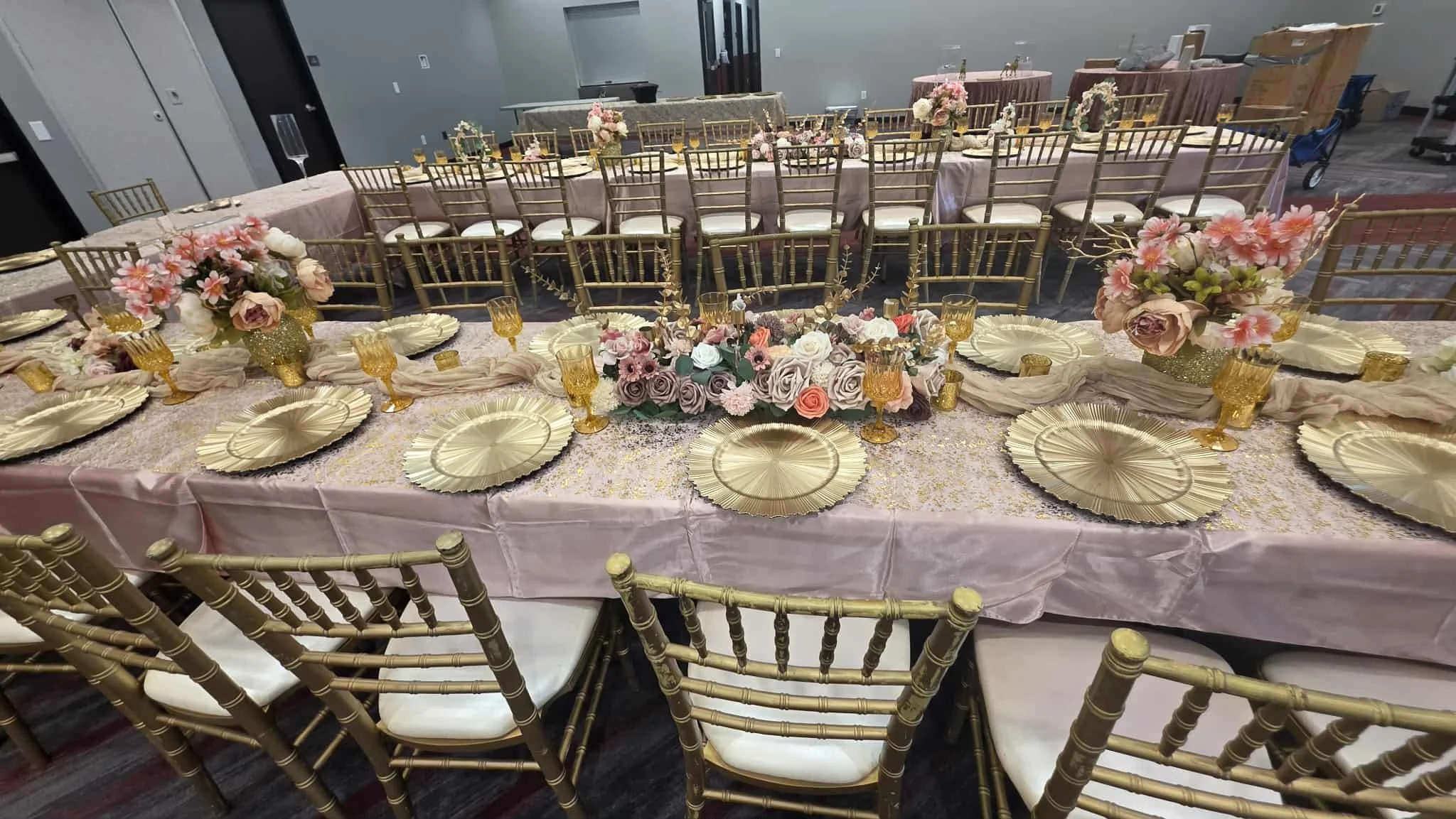 A decorated banquet table with pink tablecloths, gold charger plates, yellow glasses, and floral centerpieces with pink and white flowers and greenery, set for a formal event.