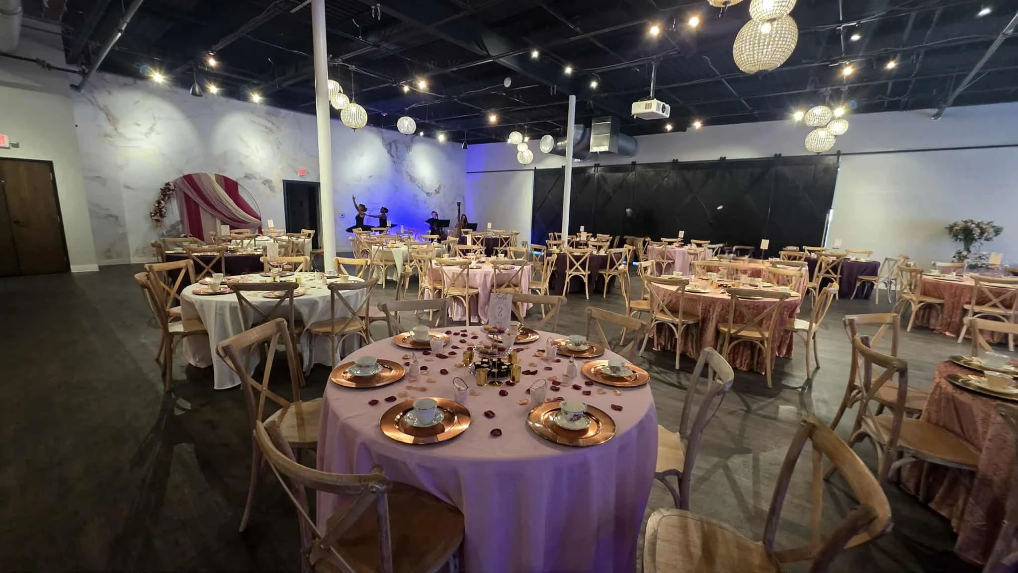 A banquet hall decorated for an event with round tables draped in pink and white tablecloths, set with plates, cups, and rose petal decorations. The room features dim lighting, chandeliers, and a performance area with dancers and blue lighting in the