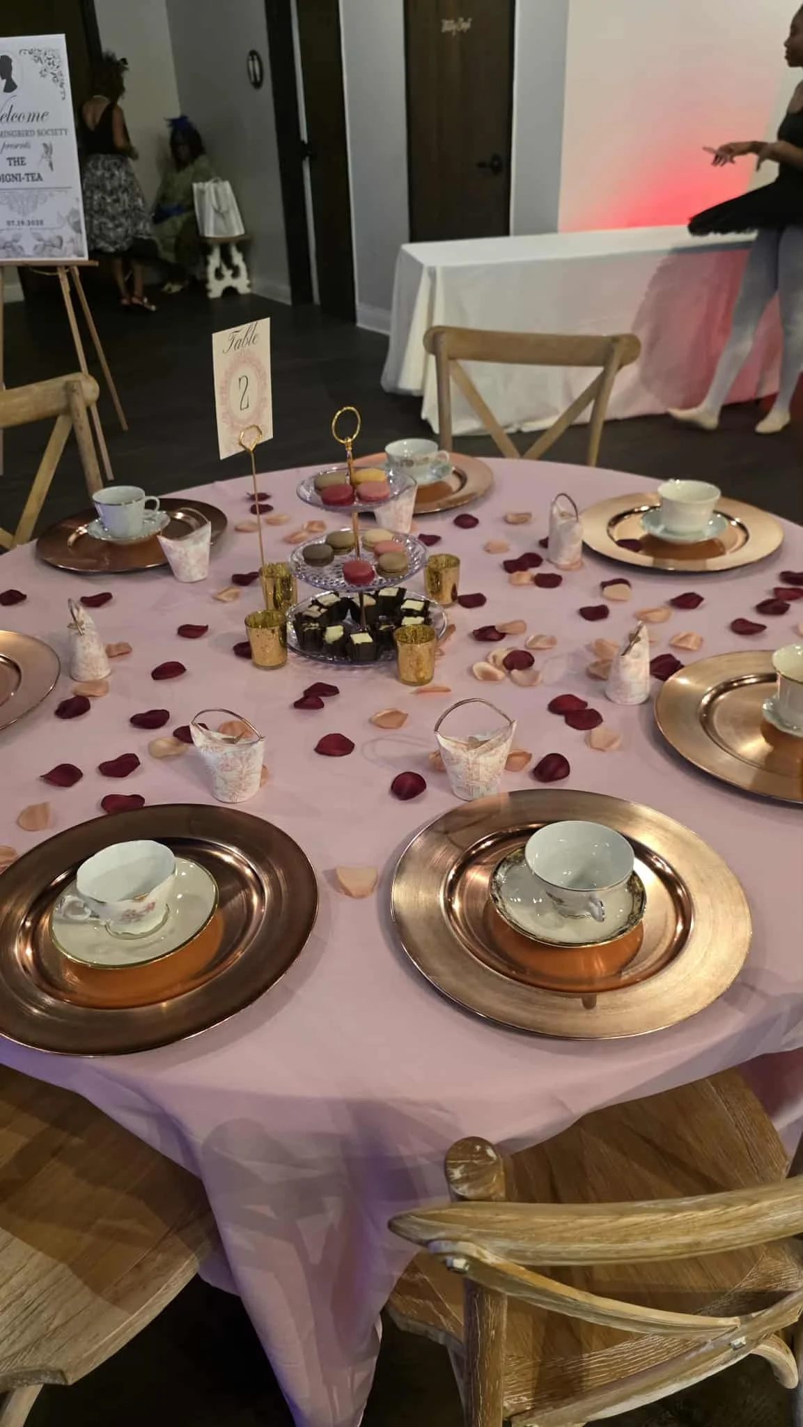 Round table with pink tablecloth set with cups and saucers on metallic gold chargers, surrounded by wooden chairs, decorated with pink and red rose petals, and central dessert stand with macarons and chocolates.