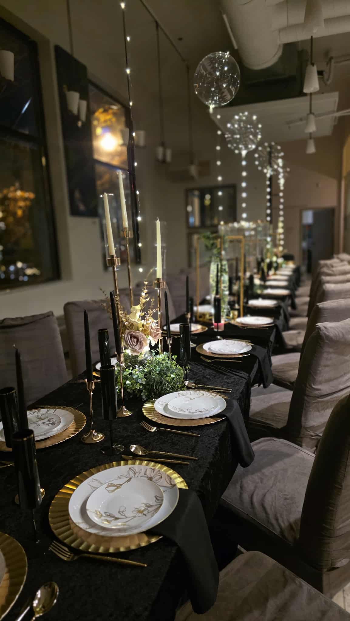 A long dining table set for a formal dinner with black tablecloth, white and gold plates, gold flatware, black and gold candlesticks with candles, and floral centerpieces. Overhead are hanging string lights and decorative globe lights, creating a festive atmosphere.