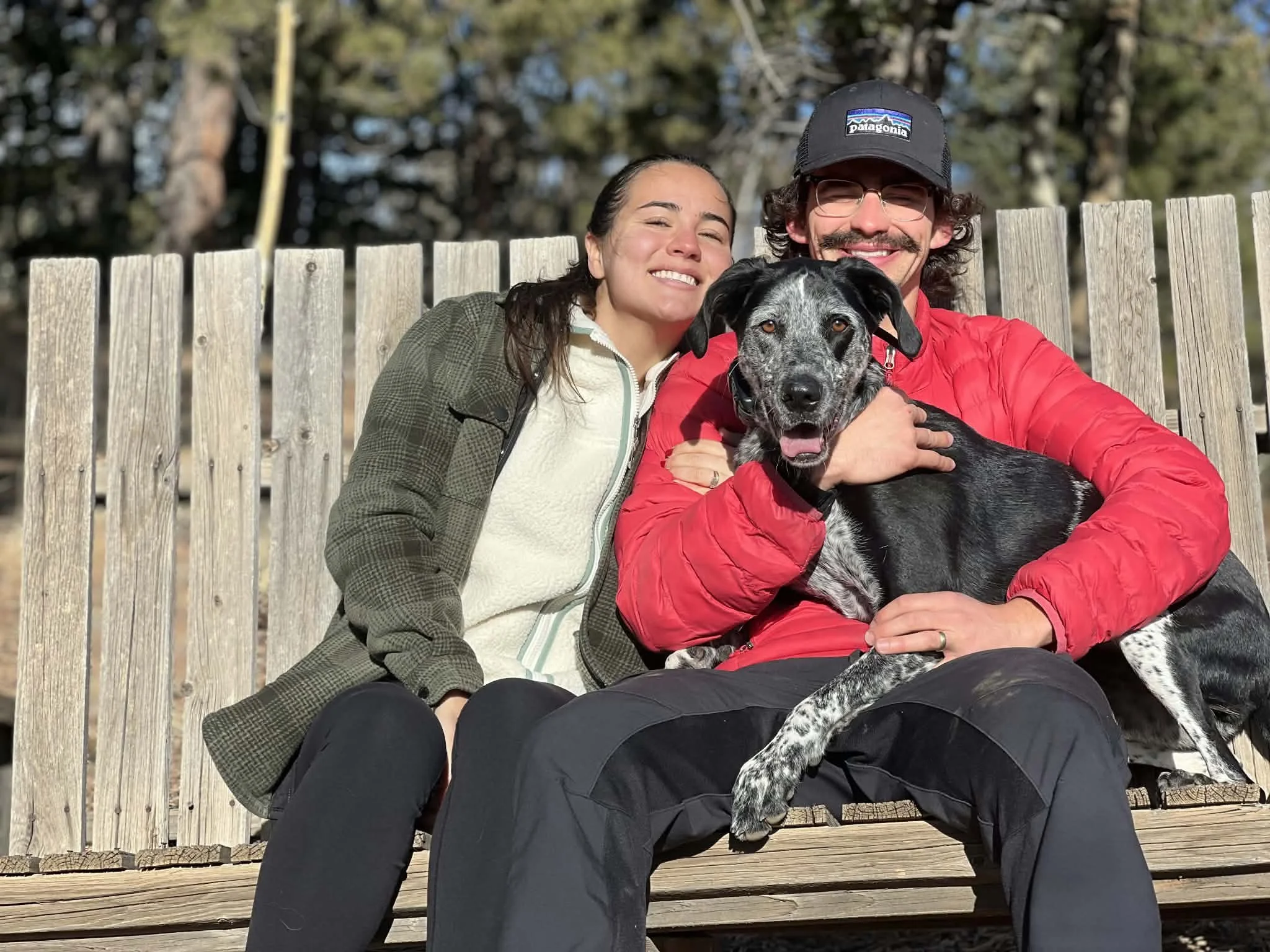 A smiling young woman and man sitting on a wooden bench outside, holding a black and white dog between them. They are dressed casually and appear happy, with a fence and trees in the background on a sunny day.