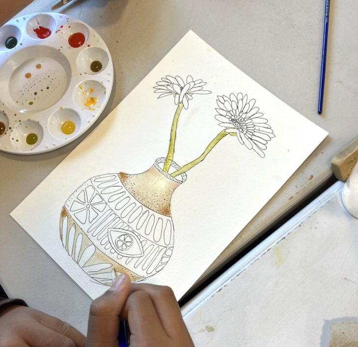 A hand drawing a vase with flowers on a white sheet of paper, with watercolor paints and a paintbrush nearby.
