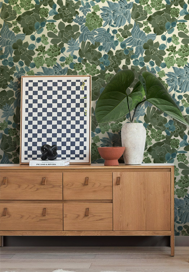 Decorative wooden sideboard with a large green-leafed plant, a terracotta bowl, a white textured vase, and a picture frame with a black sculpture and books on top, set against a botanical-patterned wallpaper.