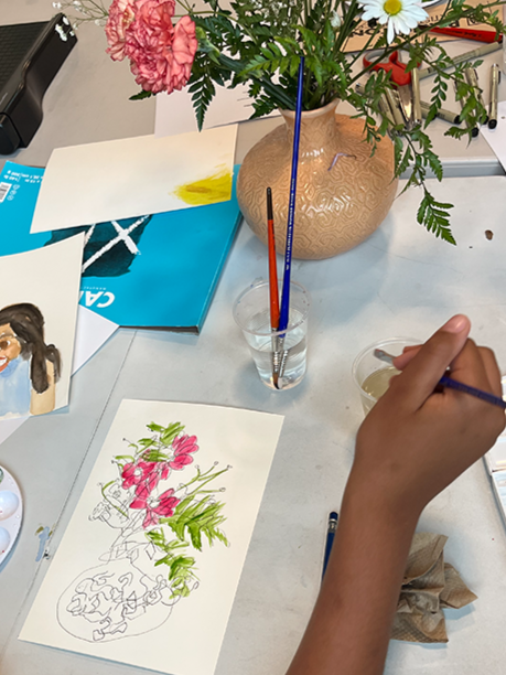 A person painting a flower arrangement on paper with a paintbrush, surrounded by art supplies, a vase of flowers, and various papers on a table.