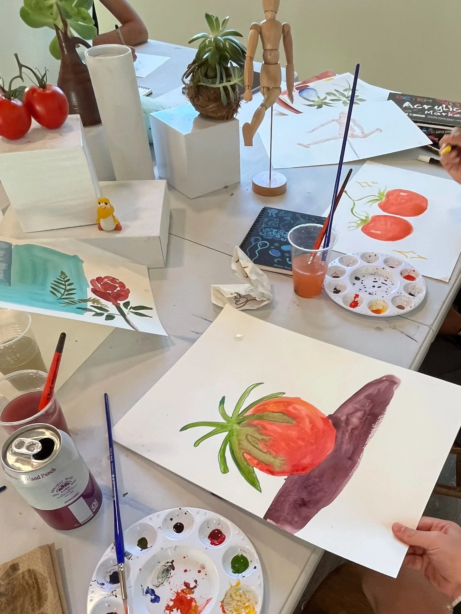 Watercolor paintings of tomatoes and a sweet potato on a table, with painting supplies including paint palettes, brushes, and water cups. People are creating art in a group setting.