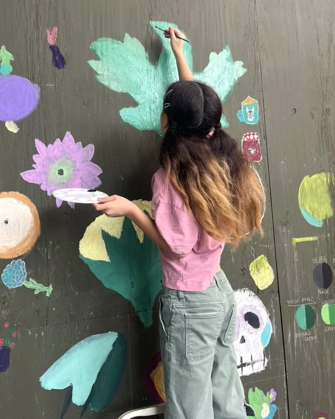 Young girl with long, wavy hair and a pink shirt using chalk to create a colorful floral mural on a wall, surrounded by other chalk drawings including a skull, fruit, and various colorful shapes.