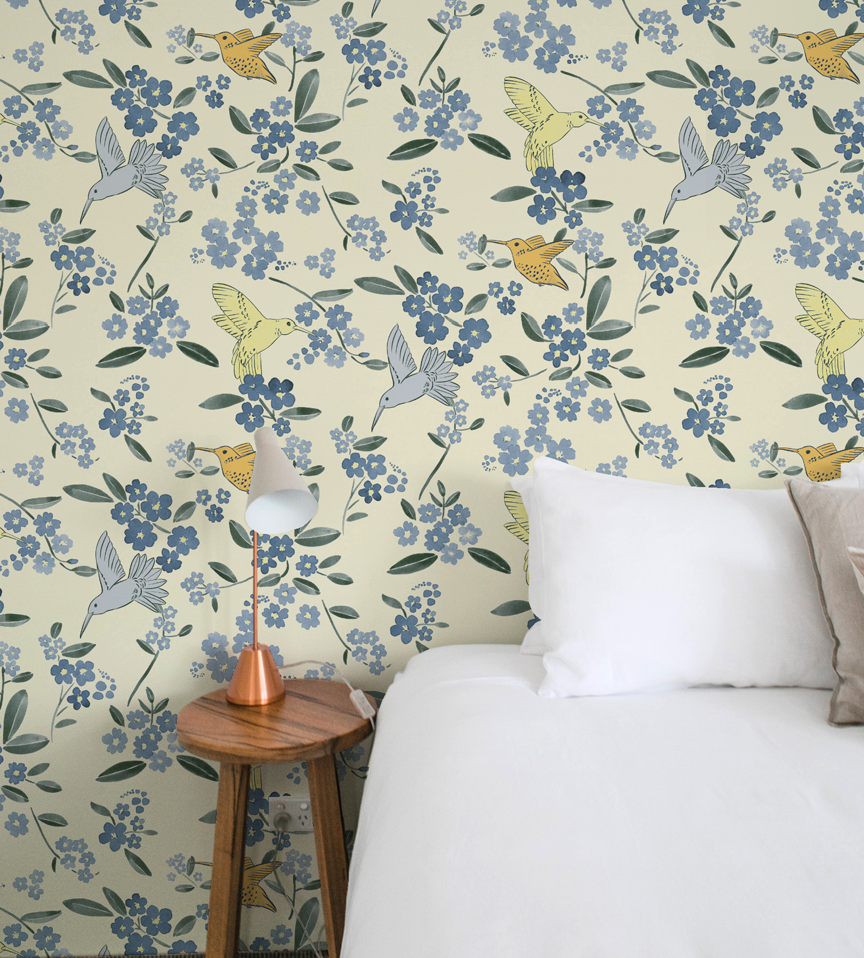 A bedroom with a wall covered in floral wallpaper with birds. A bedside table with a small lamp and a bed with white pillows.