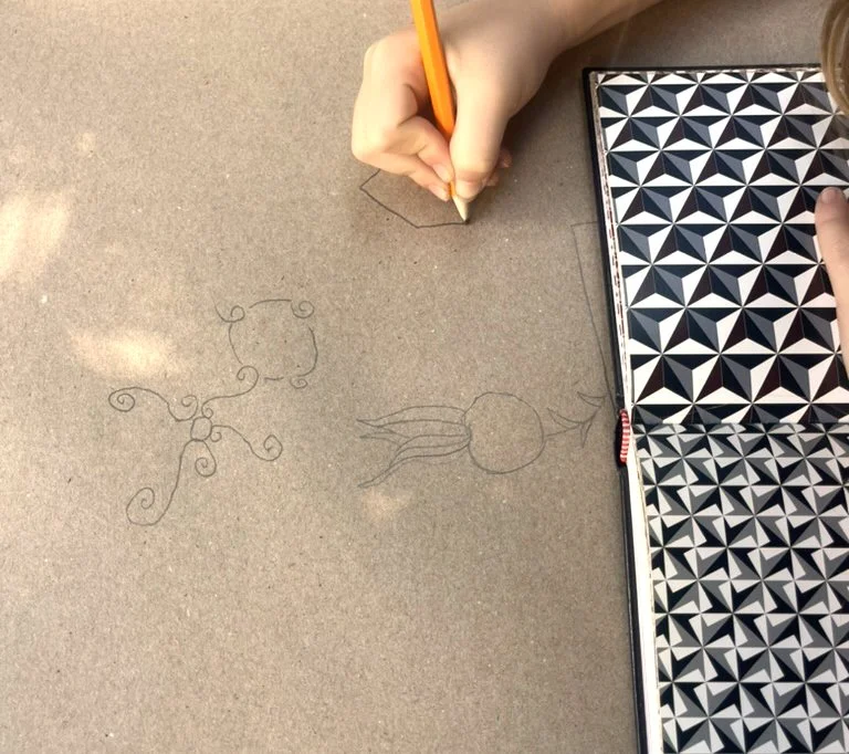 Child's hand drawing a design on brown paper with a pencil, next to a sketch of a jellyfish, and a black-and-white geometric patterned notebook.