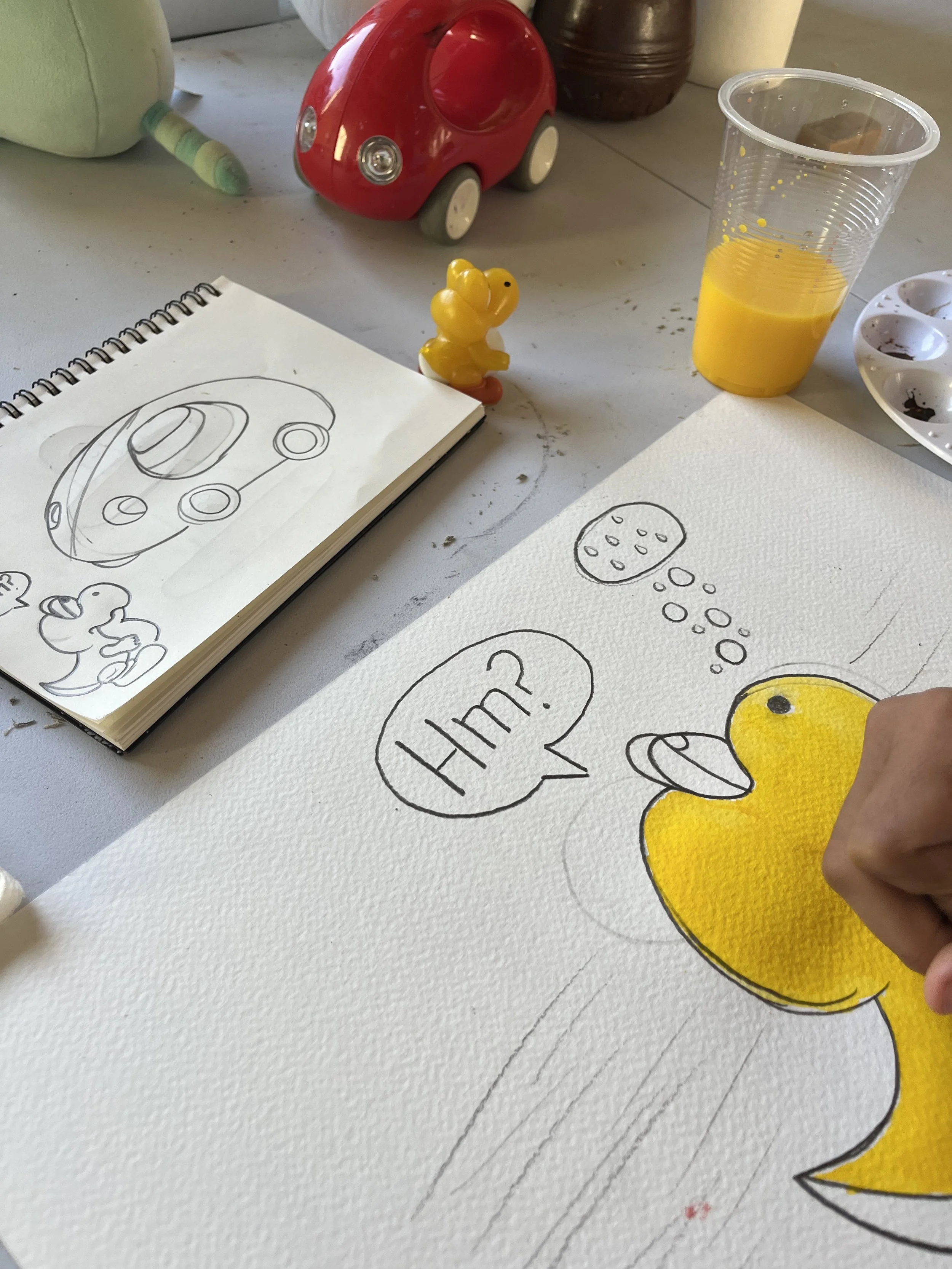 A person is drawing a yellow duck character with a speech bubble saying 'Hm?' on a piece of paper, surrounded by art supplies, a paint palette, a cup of yellow paint, a small yellow duck figurine, and children’s toys including a red toy car and a plu