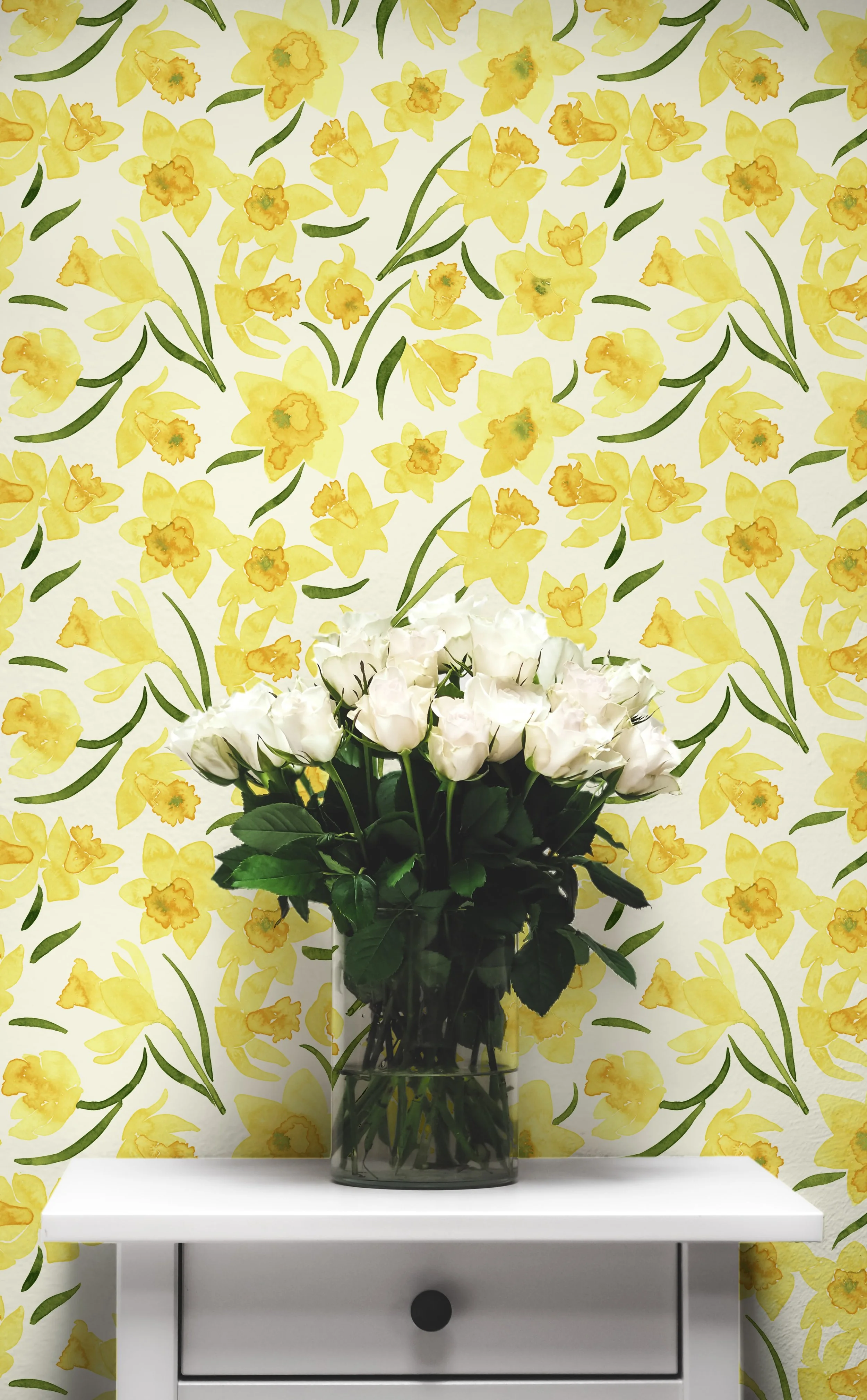 A glass vase filled with white roses, placed on a white table in front of a yellow floral wallpaper with green leaves.