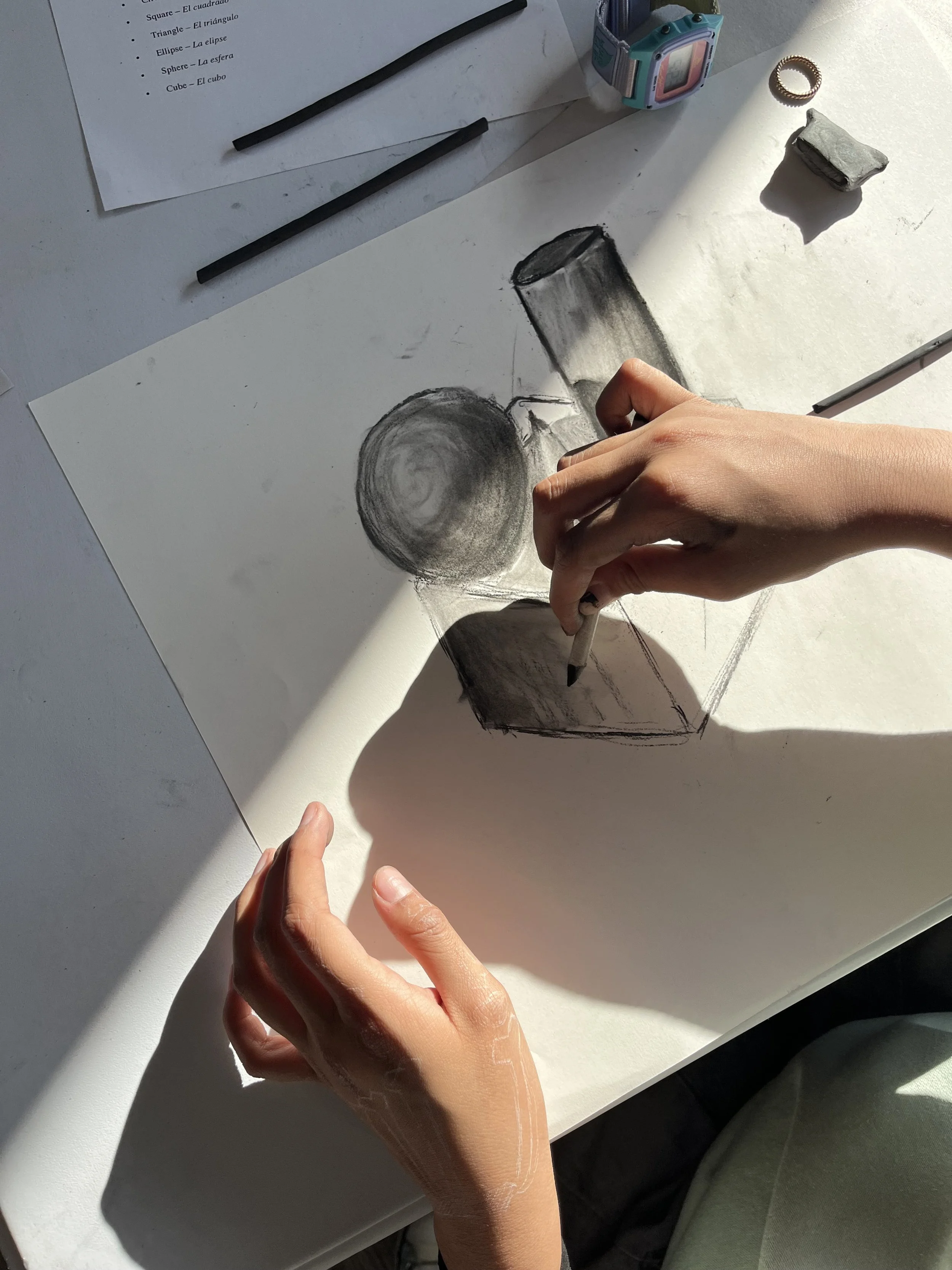 A person is drawing a still life sketch of a spherical object and a rectangular box on white paper, with sunlight casting shadows. The workspace has an eraser, a ring, a small colorful digital camera, and a sheet with geometric shape labels in Spanis
