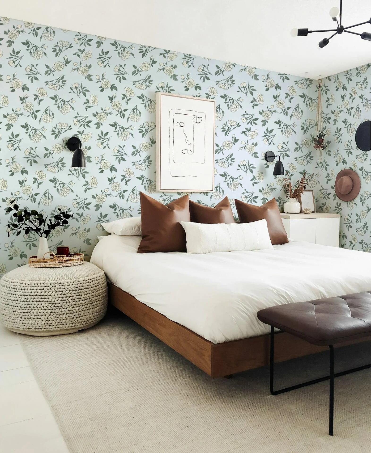 A modern bedroom with a bed that has white bedding and brown pillows, a woven pouf at the foot of the bed, a cream-colored rug, a white dresser with decor, and wall-mounted black lamps on floral wallpaper background.