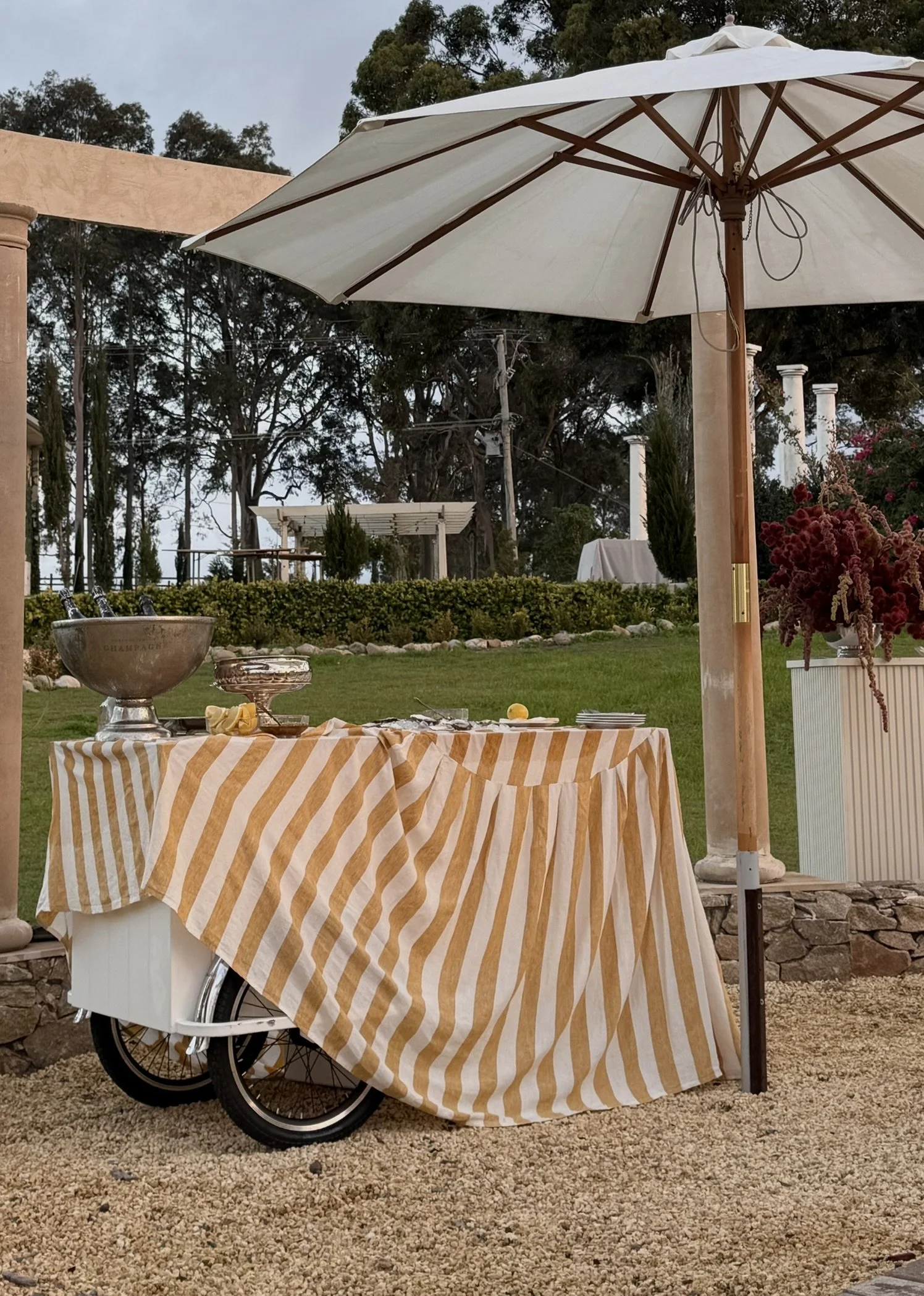 A food cart with a yellow and white striped tablecloth, adorned with oysters on ice, a champagne bowl with champagne and cavir. A large white umbrella over it, set in an outdoor garden area with trees and columns in the background.