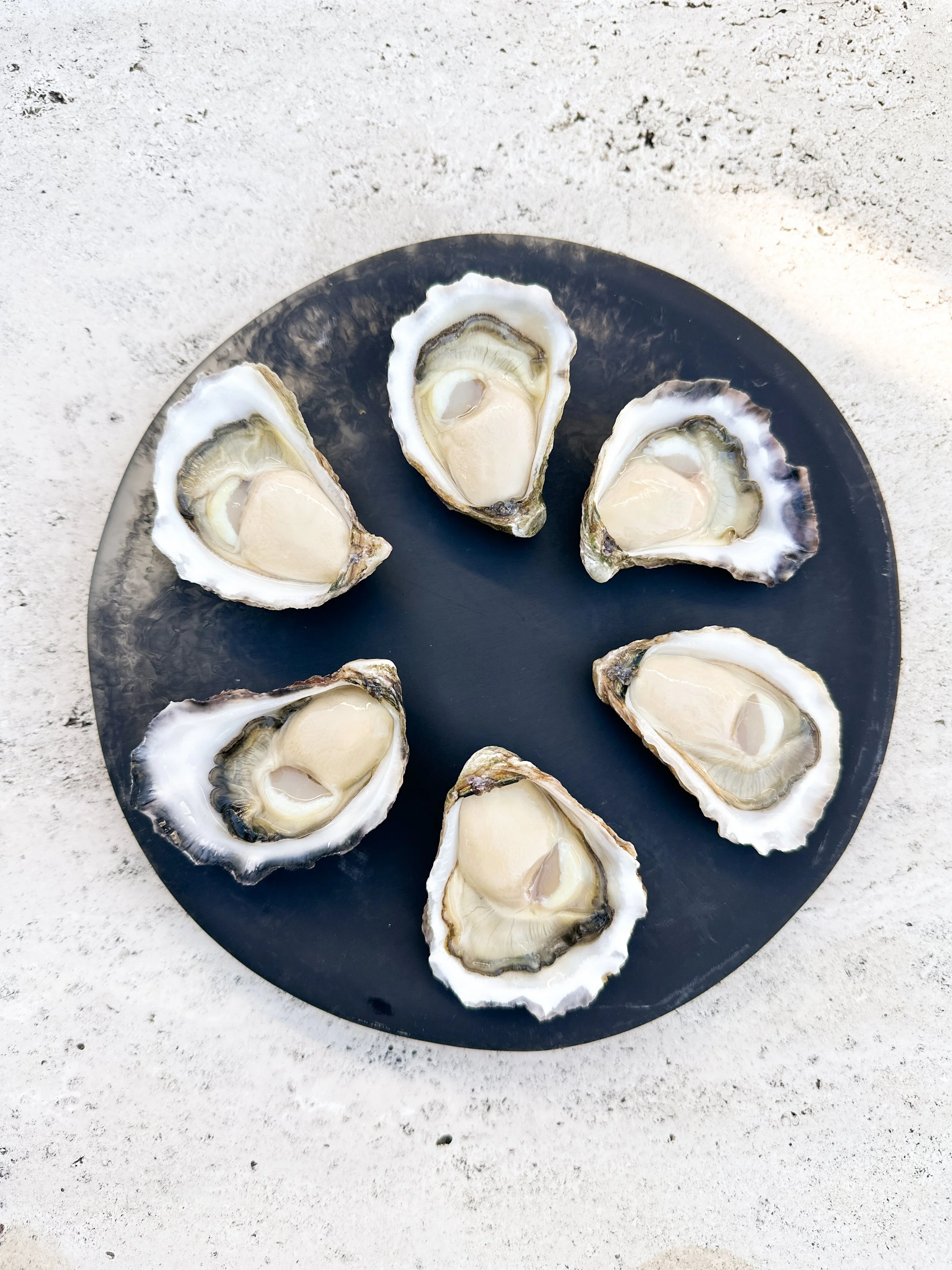 Six oysters on a black plate, set on a white textured surface.