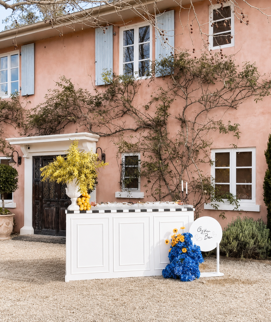 A white oyster bar stand decorated with yellow and blue flowers, placed in front of a pink house with blue shutters, outdoor plants, and leafless vines.